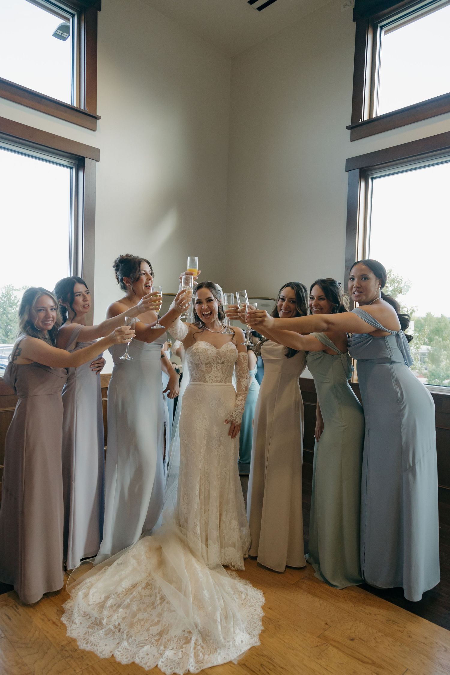 Bride in lace gown toasting champagne with bridesmaids in blue and gray dresses by tall windows.