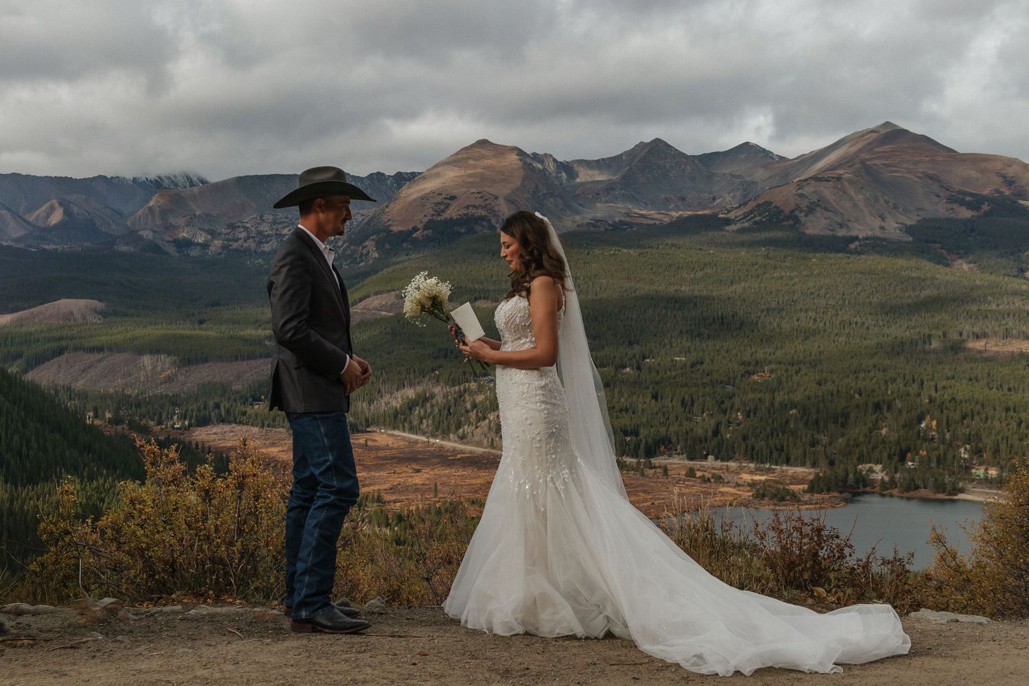A couple stands on a mountain overlook with sweeping views of peaks and a lake in the Colorado wilderness.