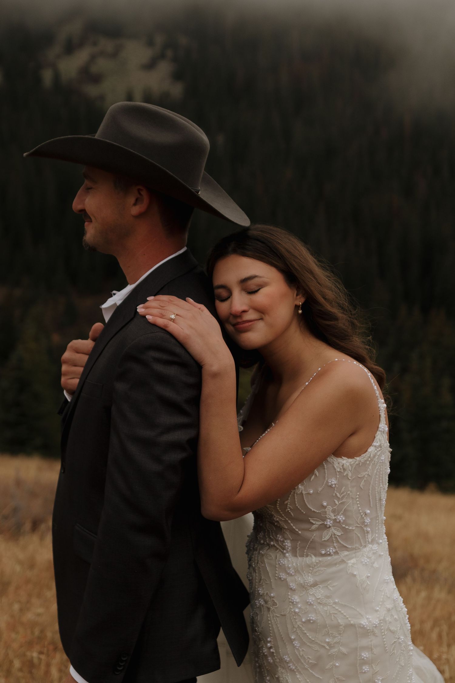 A couple in wedding attire embraces in a golden field with mountains in the background.
