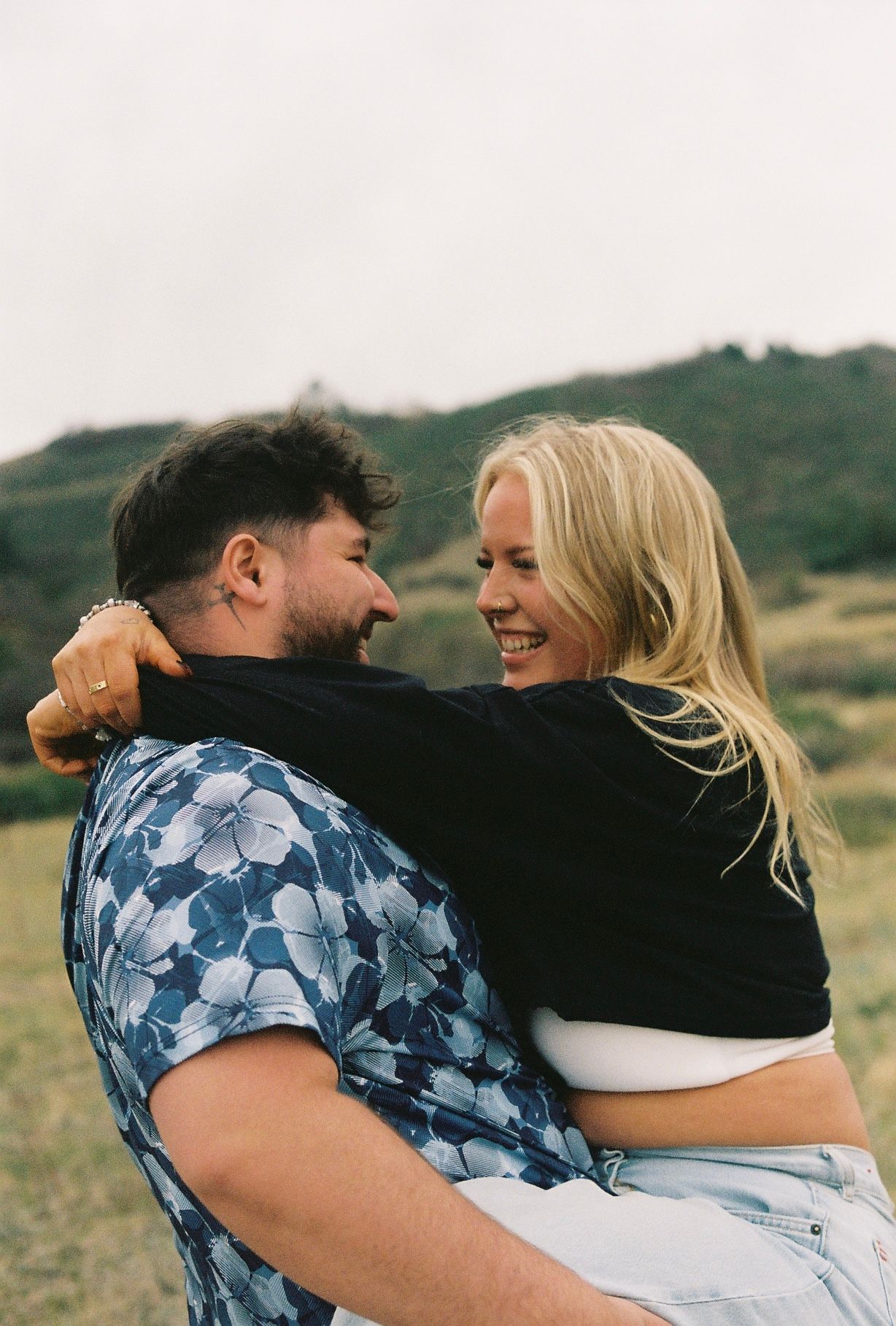 A smiling blonde woman embraces a bearded man in a floral shirt outdoors with green hills in the background.