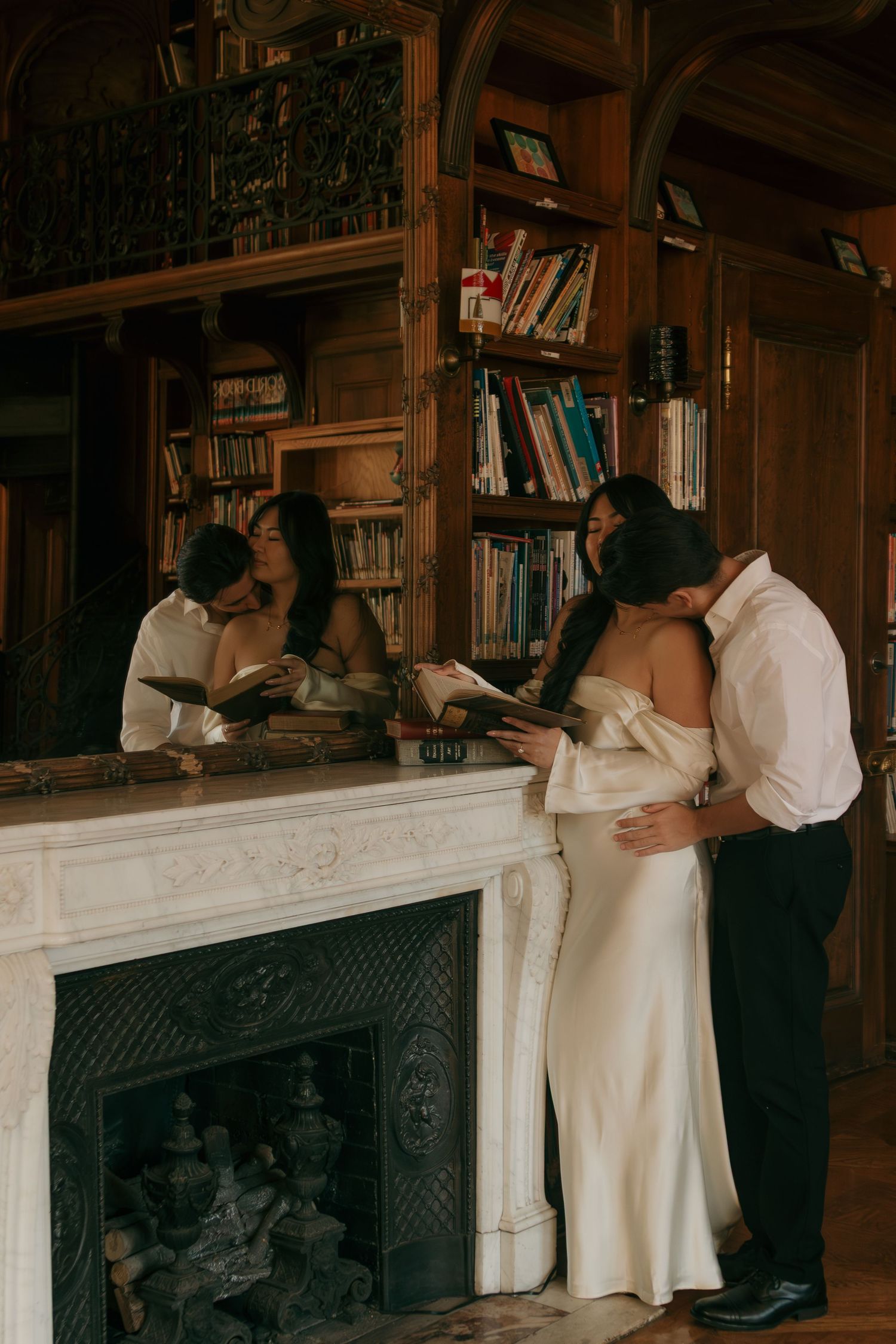 Couple in wedding attire standing by an elegant marble fireplace in a grand library setting.