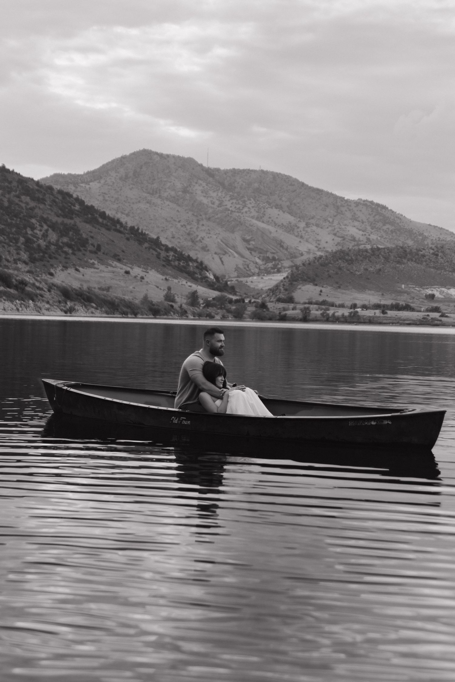 Black and white photo of a canoe on calm lake waters with mountains in the background.