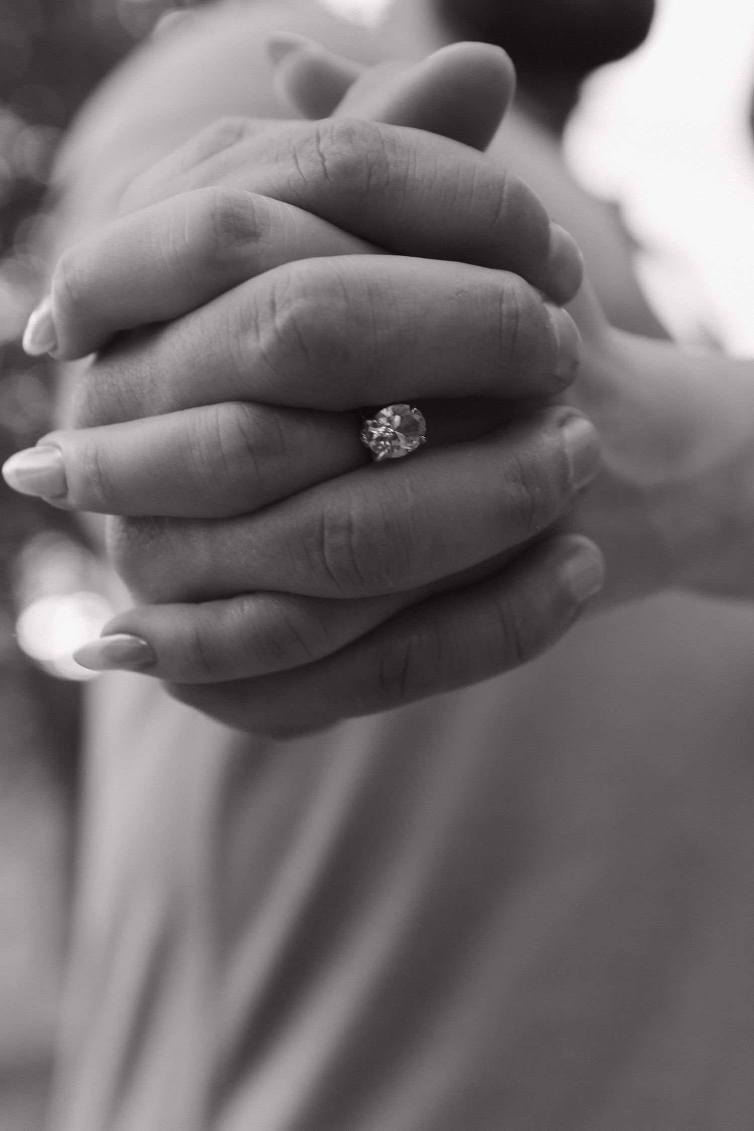 Close-up black and white photo of intertwined hands with an engagement ring.