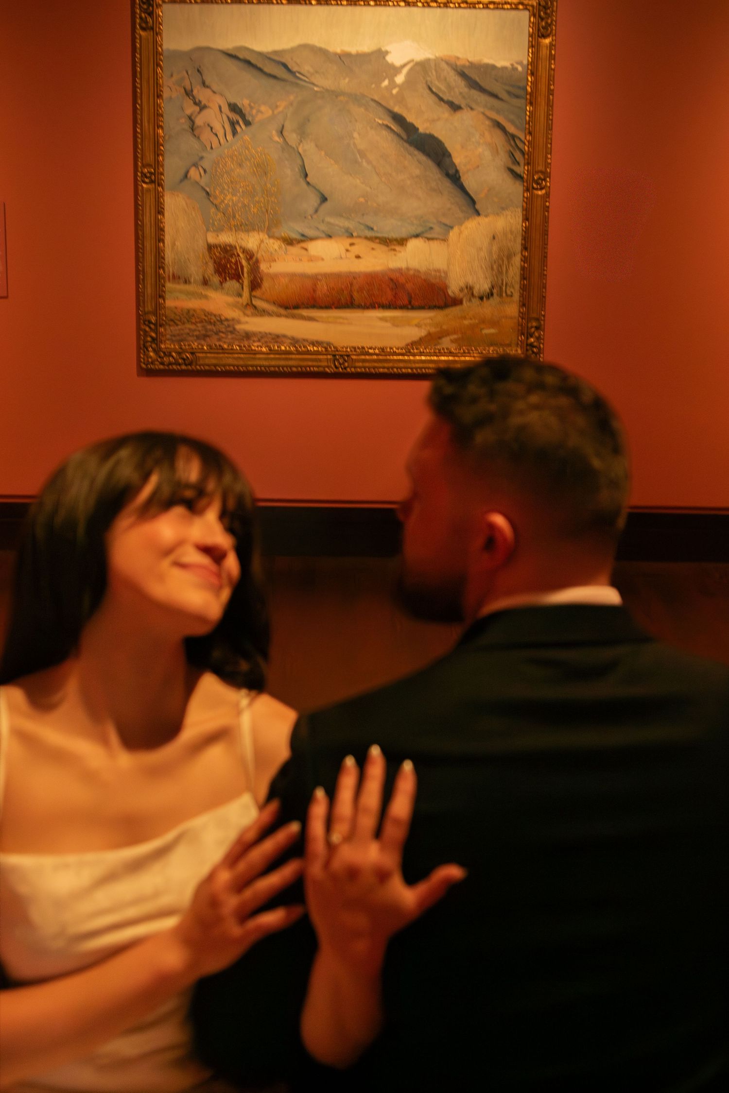Romantic couple sharing an intimate moment in an art gallery with landscape paintings on terracotta walls.
