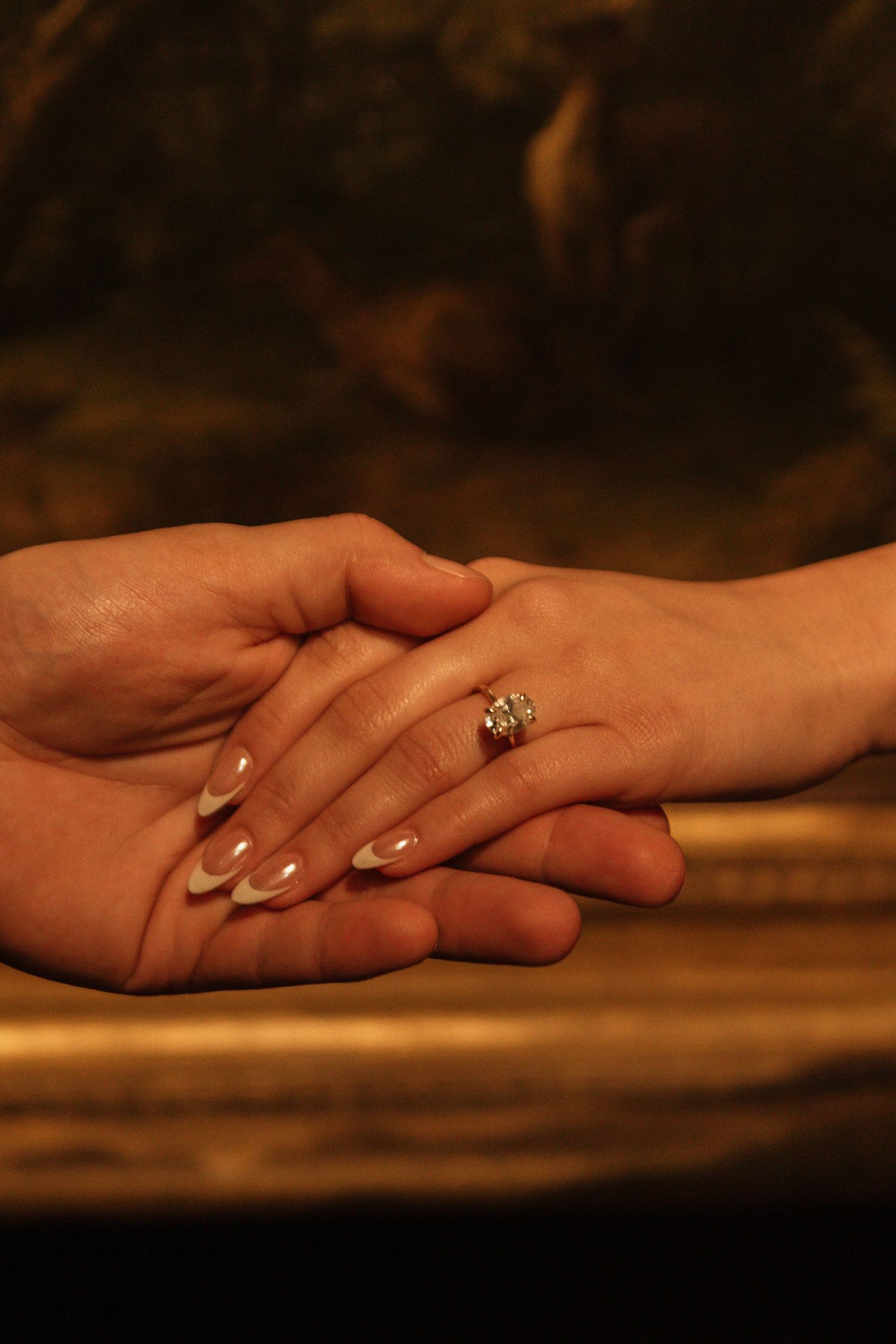 Close-up of two hands clasped together with an engagement ring visible in warm lighting.