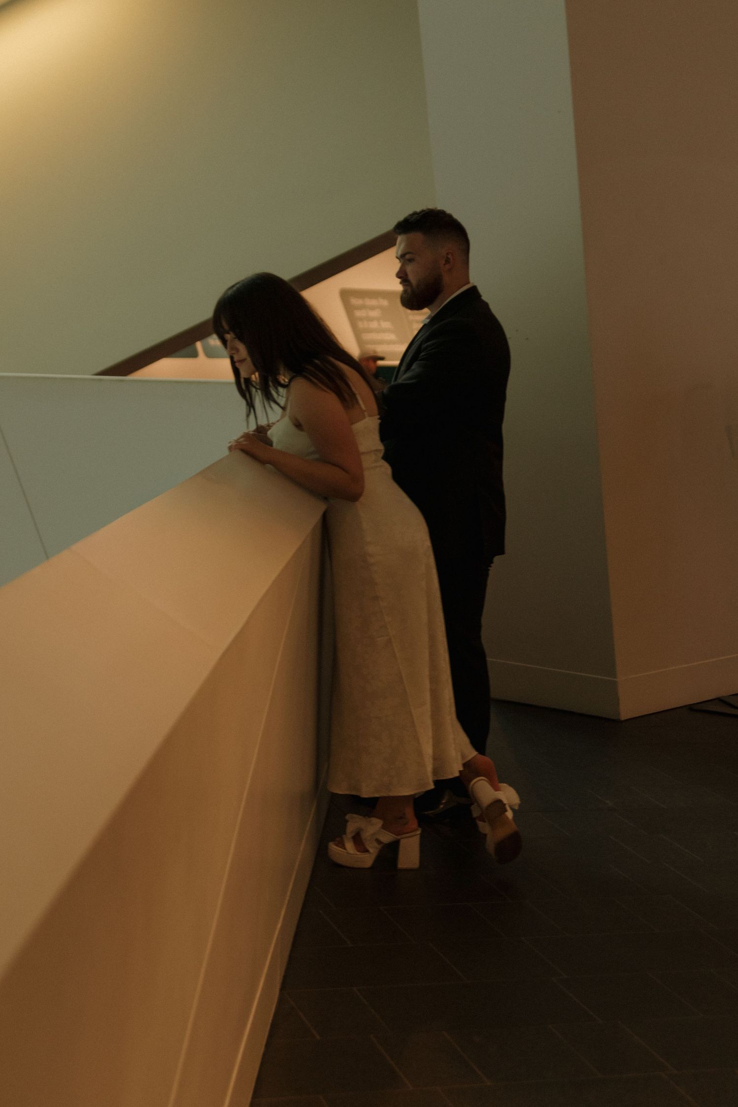 Two figures stand at a white gallery wall examining artwork together in soft lighting.