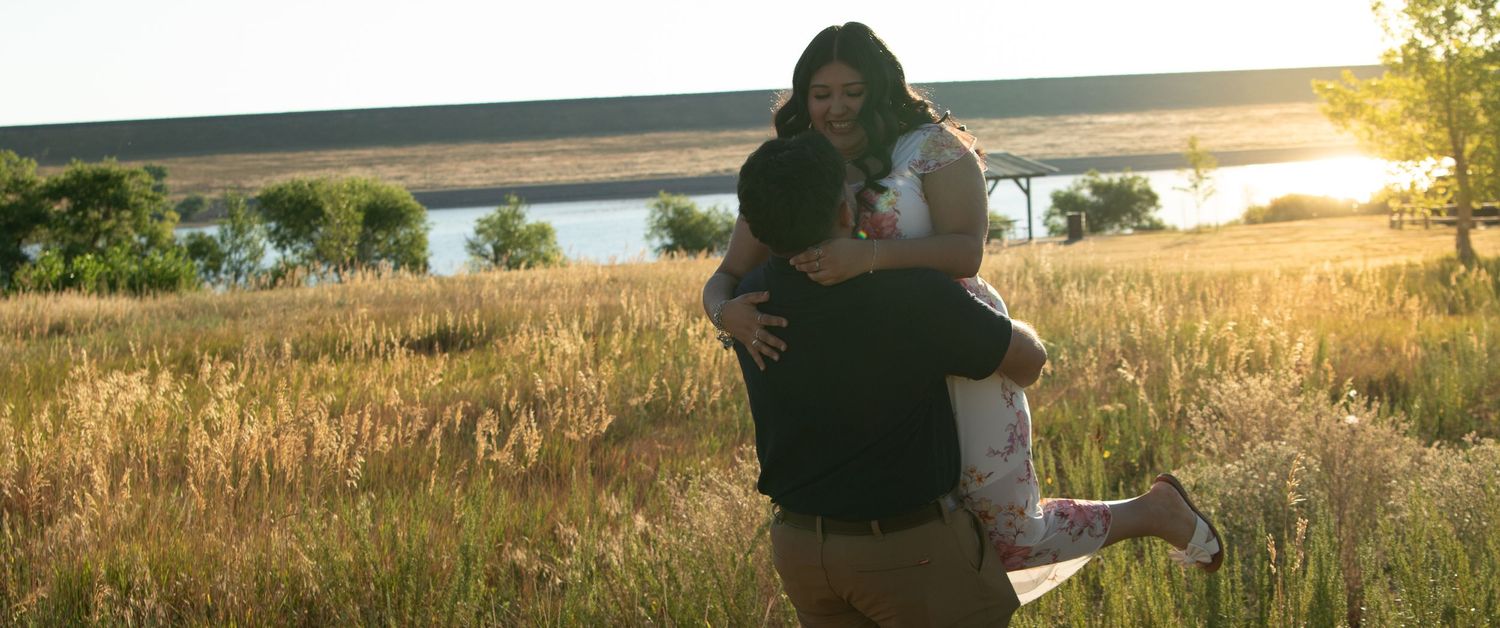 A couple shares a tender moment at sunset near a lake with tall grass surrounding them.