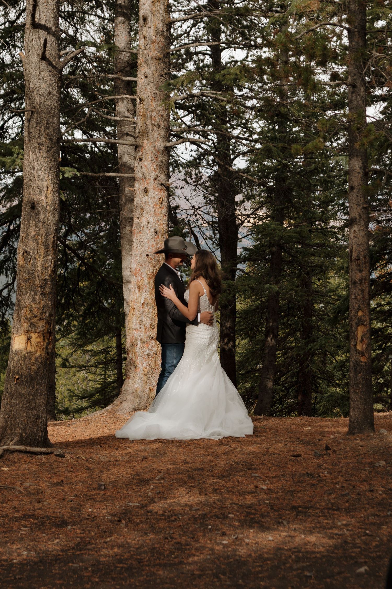 A wedding couple shares an intimate moment among tall pine trees on a forest trail.