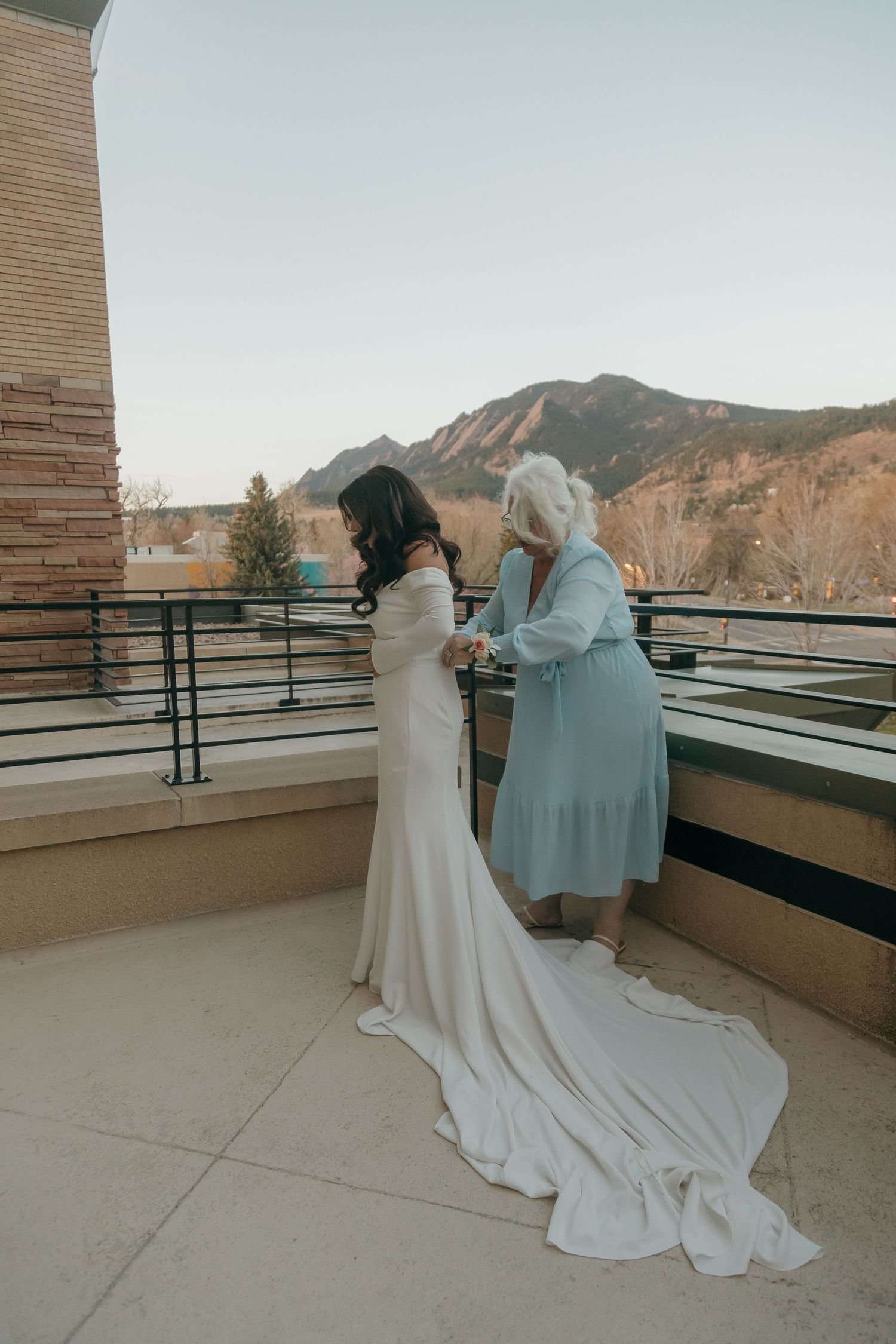 Bride in white gown with train stands on balcony with elderly woman, Flatirons mountains in background.