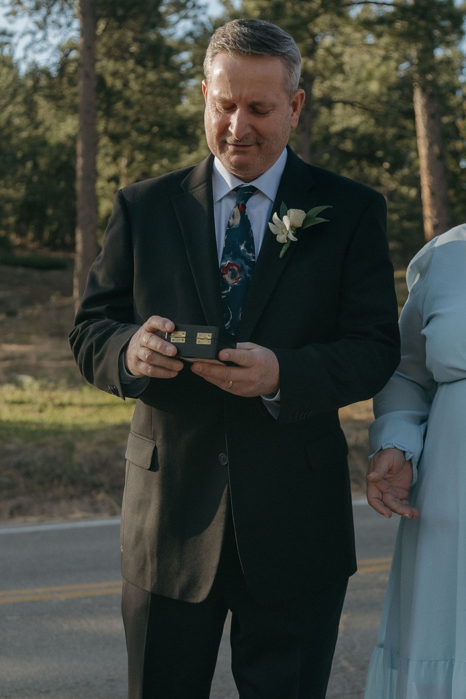 Groom in black suit smiles while holding wedding ring box during outdoor ceremony.