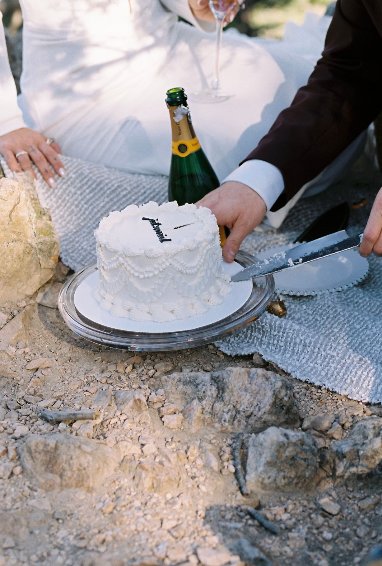 Bride and groom cut a small white wedding cake beside a champagne bottle on a rocky outdoor surface.