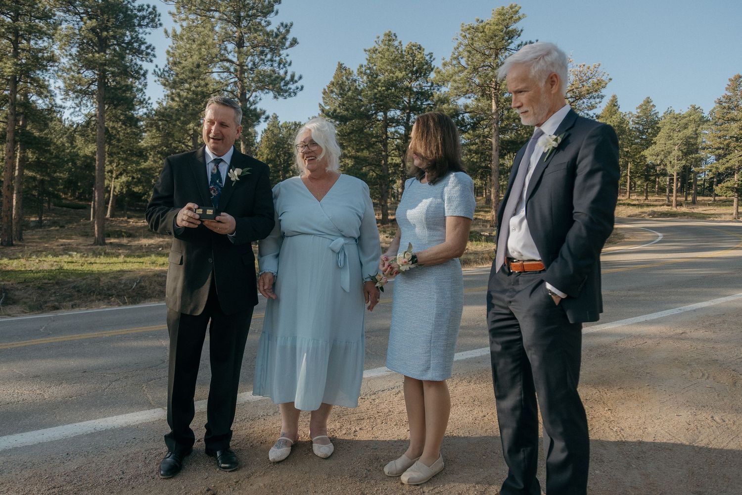 Four wedding guests in formal attire gather and converse on a sunlit mountain road after ceremony.