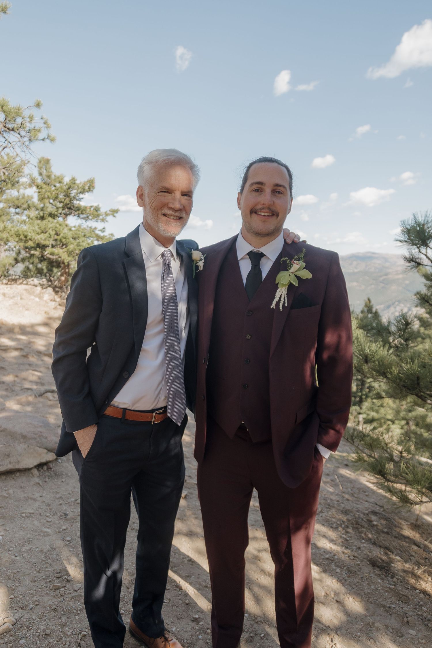 Groom in burgundy suit smiling with older man in dark suit at outdoor mountain wedding.