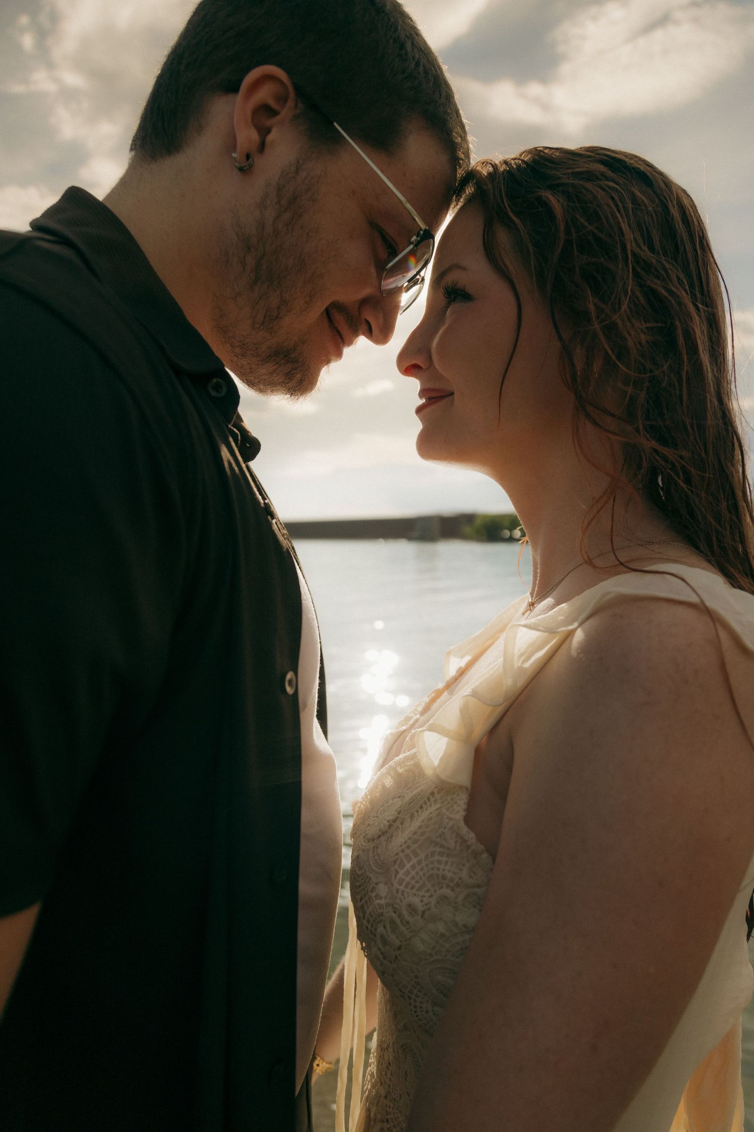 Romantic couple touching foreheads intimately with a sunlit lake backdrop.
