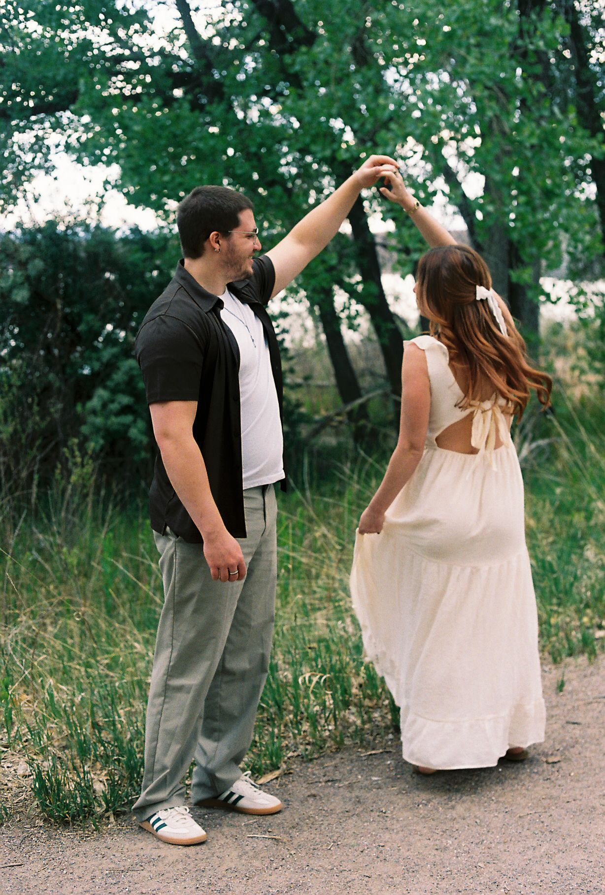 Man spinning woman in white dress on a dirt path surrounded by lush green trees.