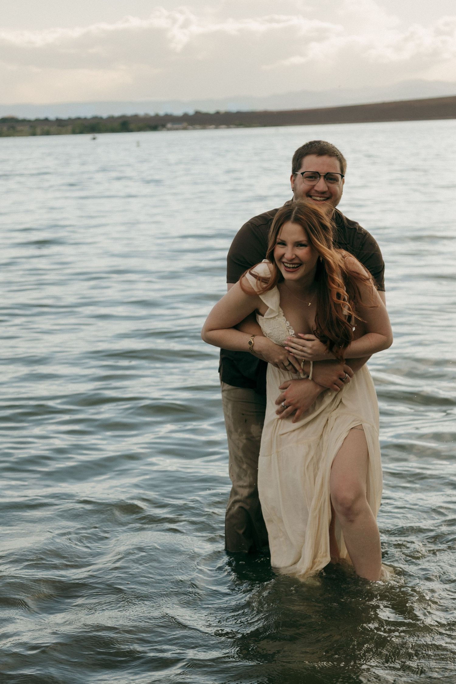 Couple laughing and embracing while standing in a lake at sunset, woman in cream dress, man in black shirt.