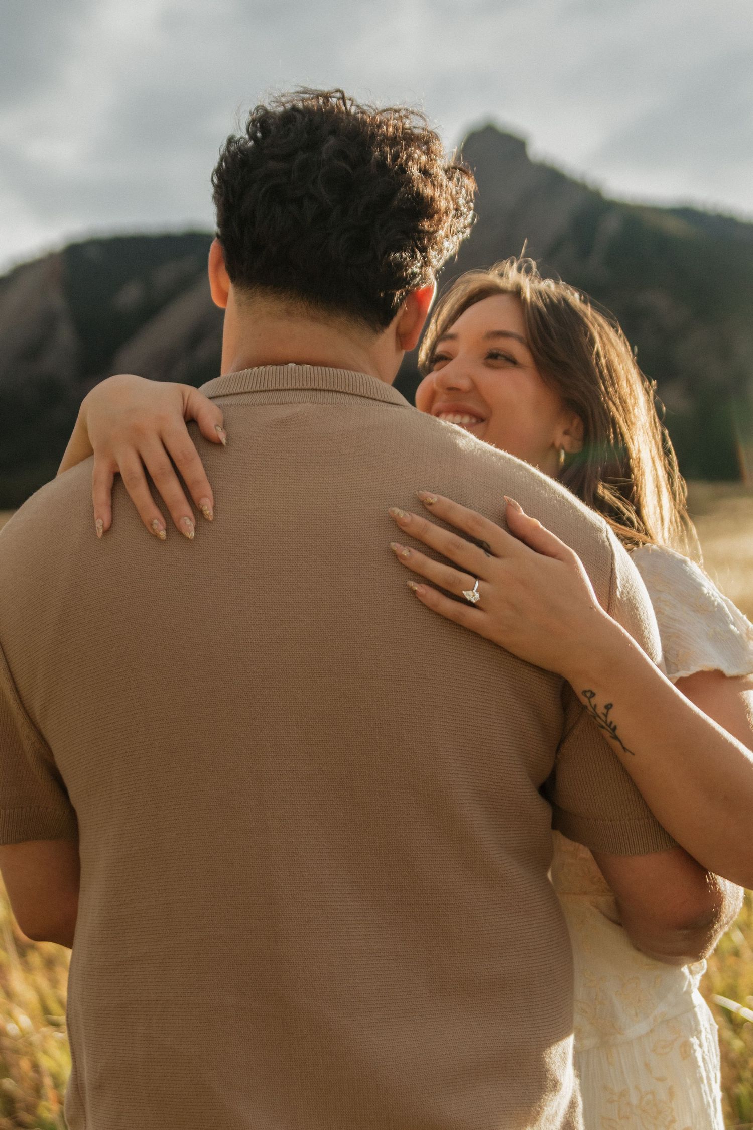 Woman in white dress hugging man from behind showing engagement ring with rocky mountains backdrop.