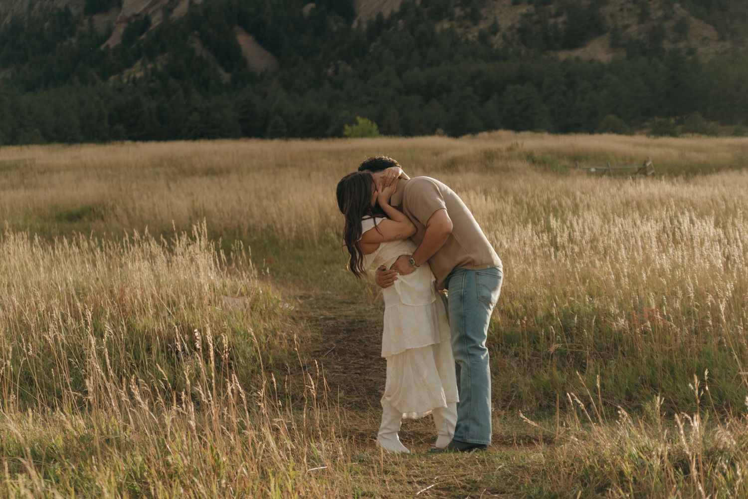 Couple shares a deep romantic dip kiss on a sunlit path through tall golden Colorado meadow grass.