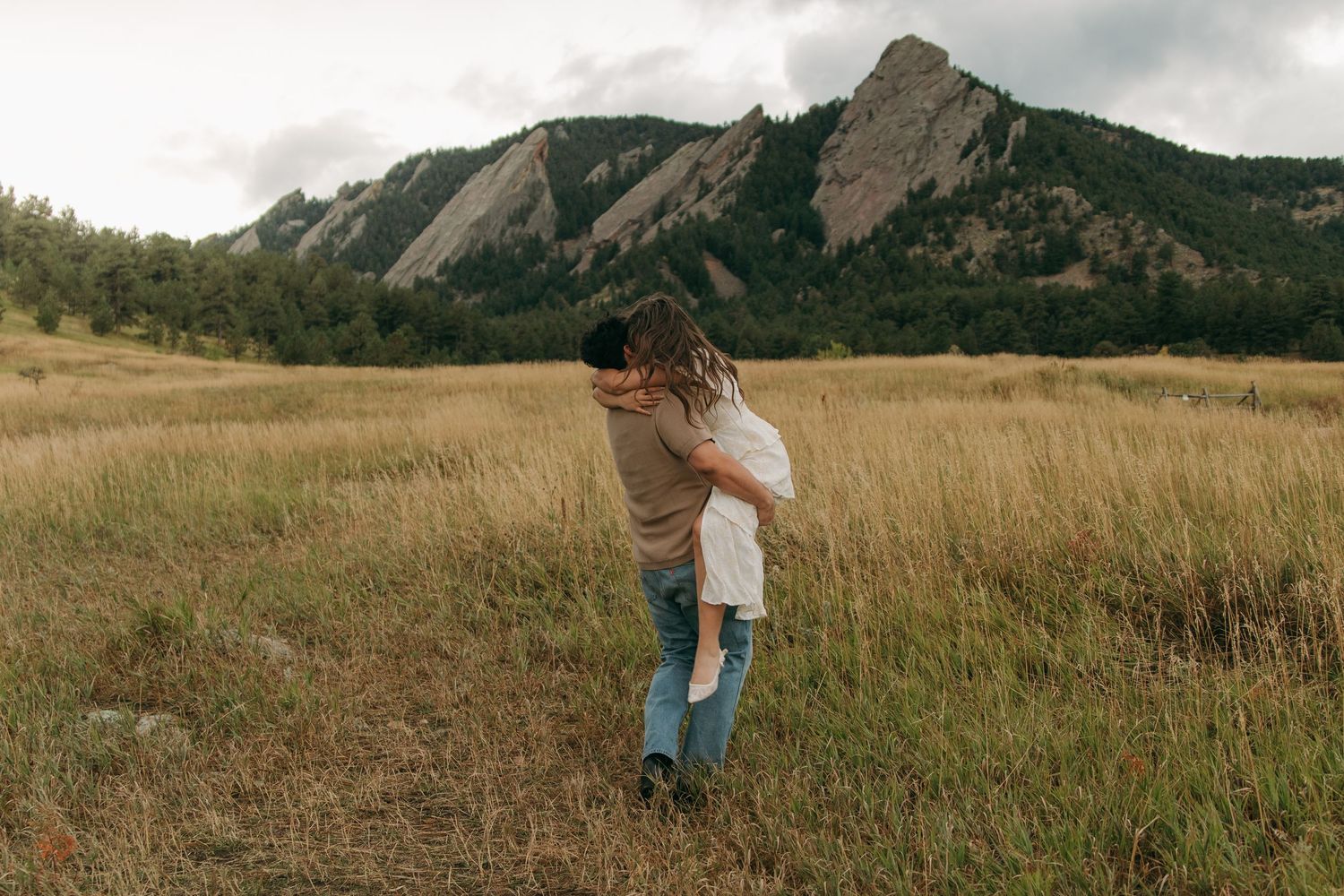 Couple embracing in golden meadow field with iconic Flatirons mountain formation behind them.