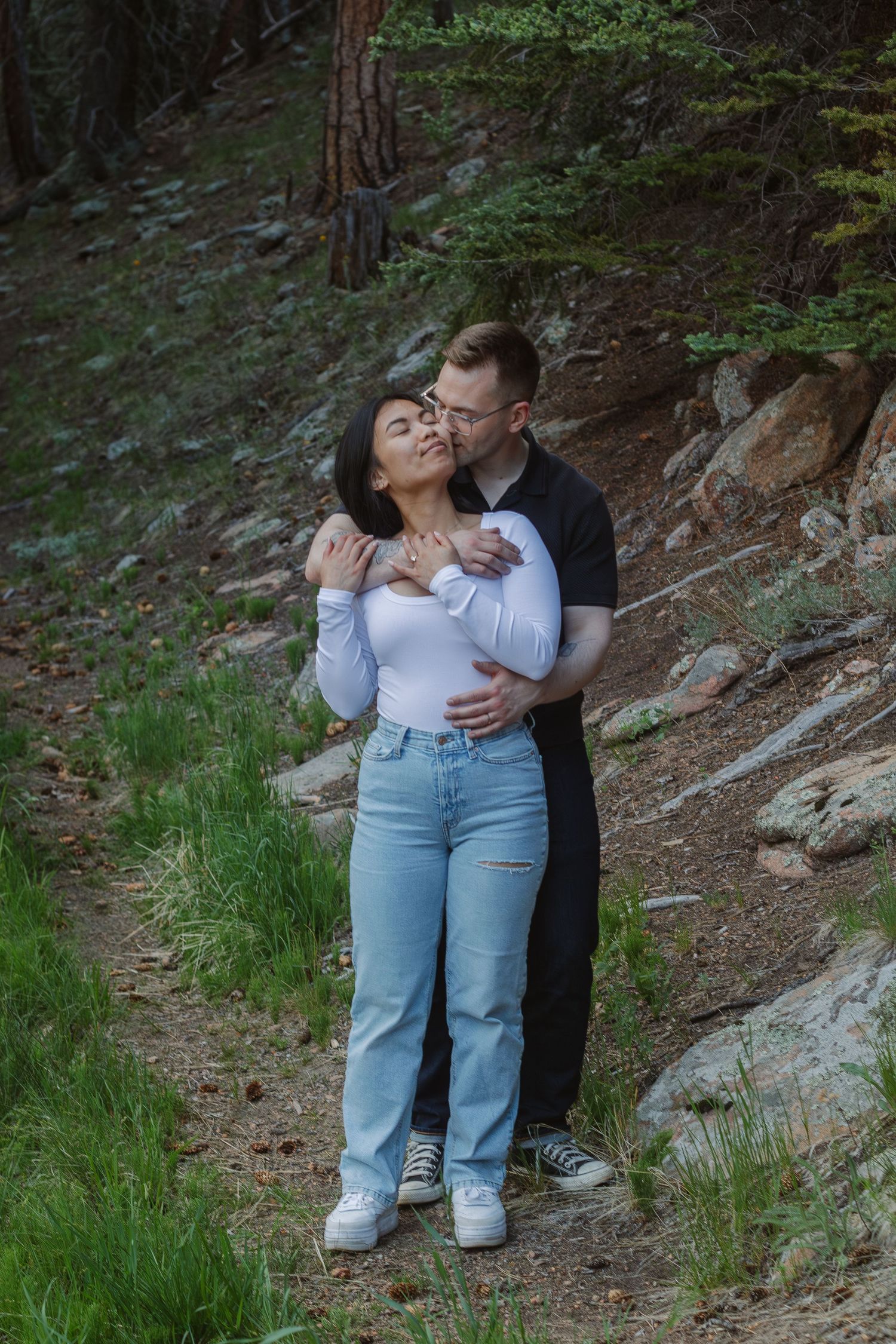 A couple shares an intimate embrace while standing on a forest trail surrounded by evergreens.