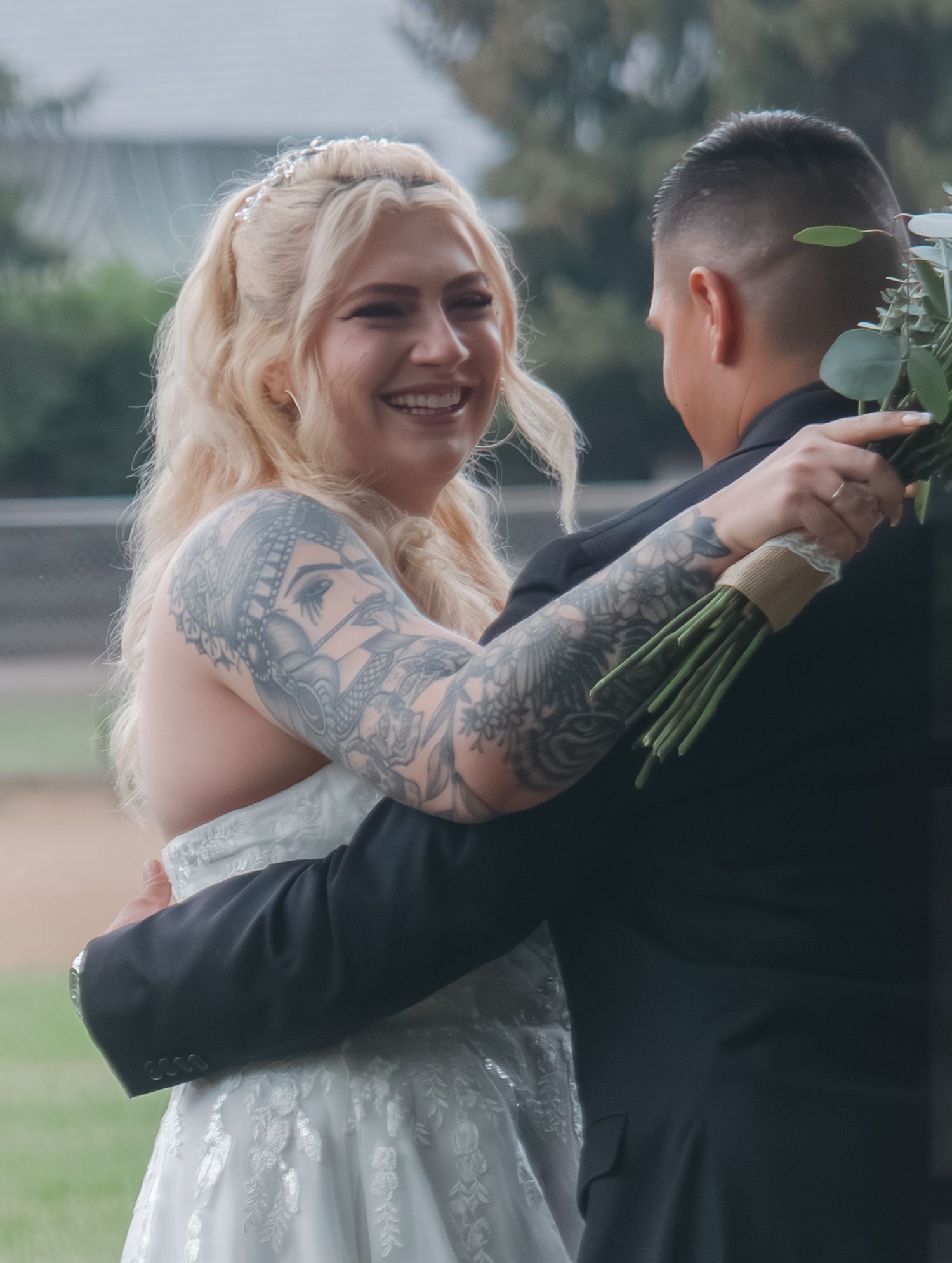 A bride with blonde hair and tattoos embraces someone while holding a bouquet outdoors.