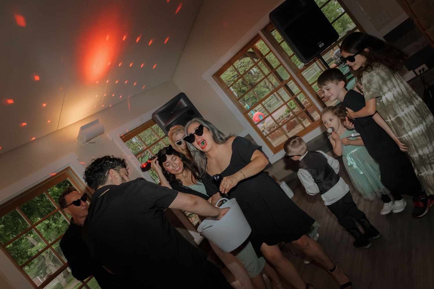 A group dances together at a party under moody red lighting and windows.