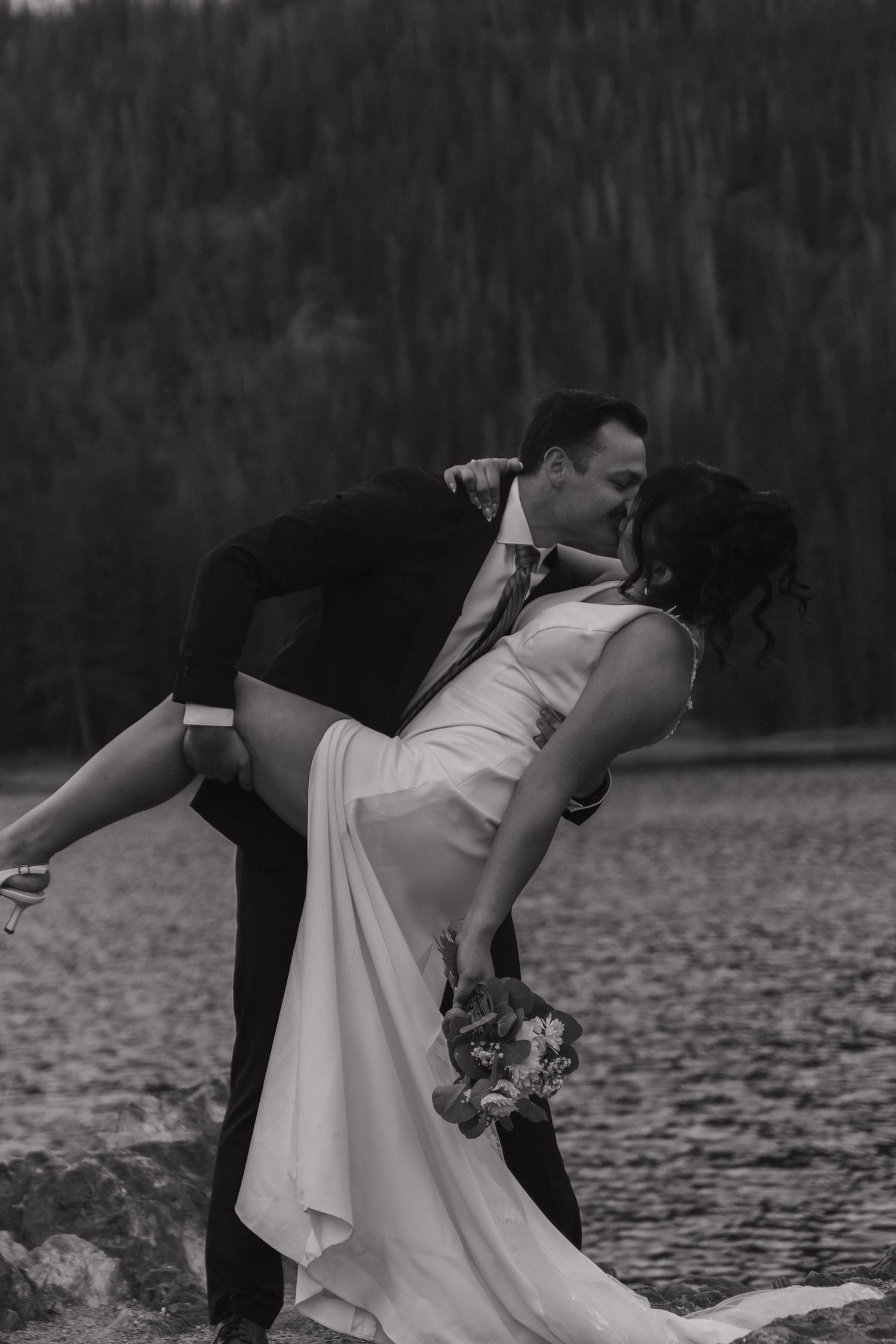 Romantic black and white dance dip pose by lake with mountains in background.