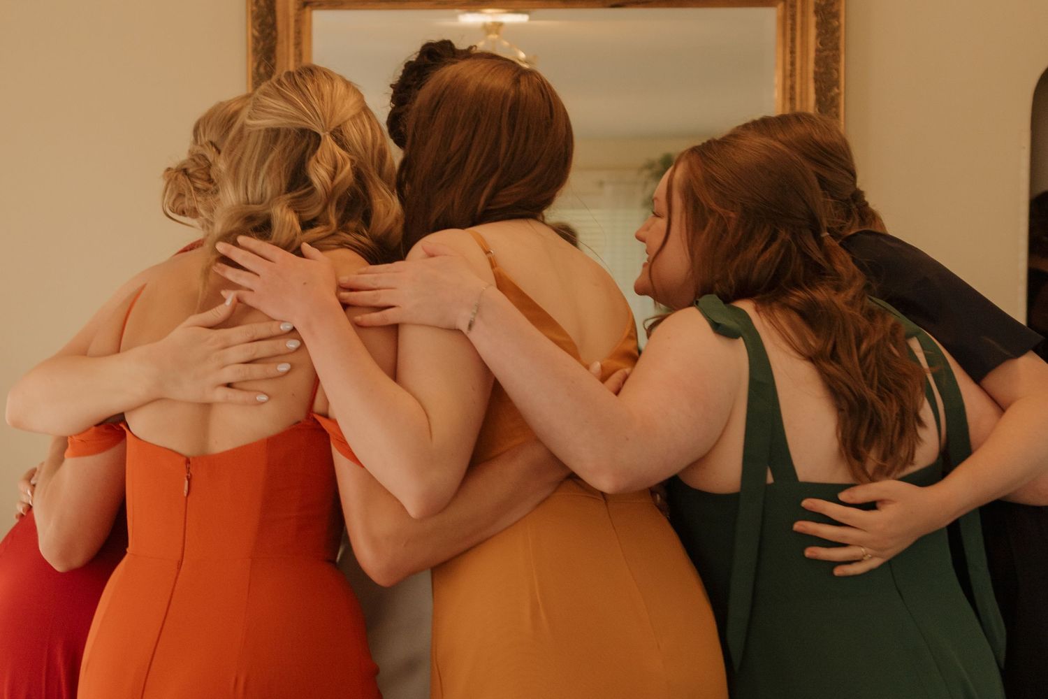Bridesmaids share an emotional embrace in a group hug wearing red and orange dresses.