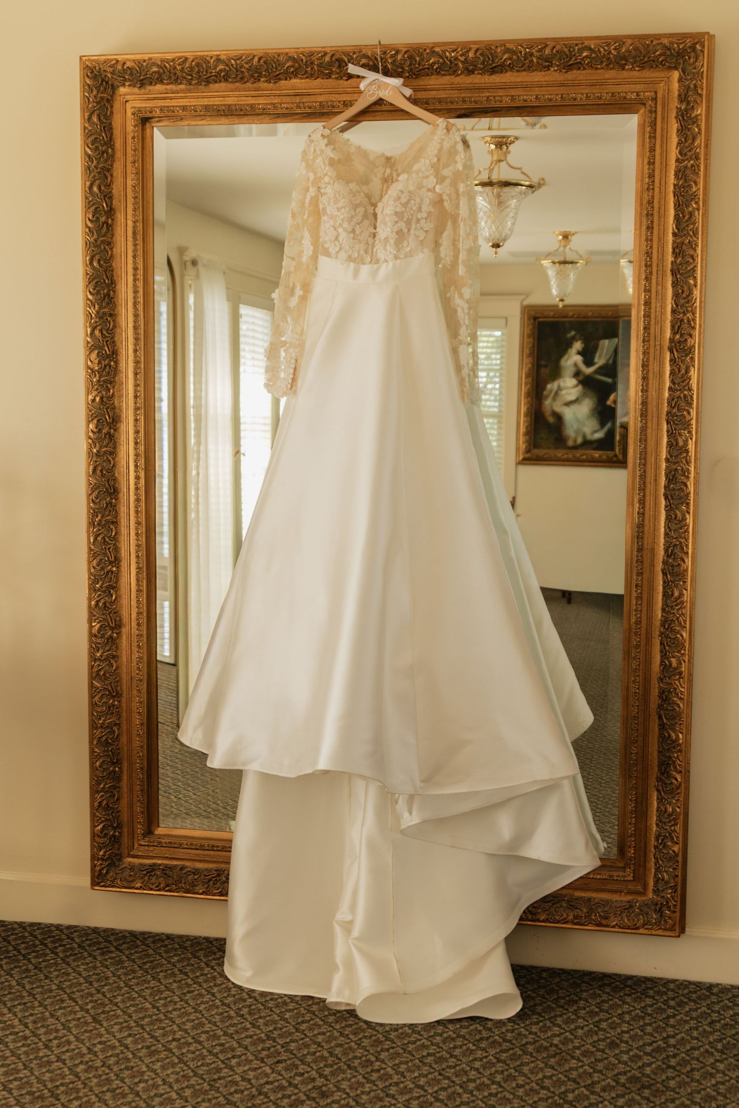 An elegant white wedding gown hangs in front of an ornate gold-framed mirror.
