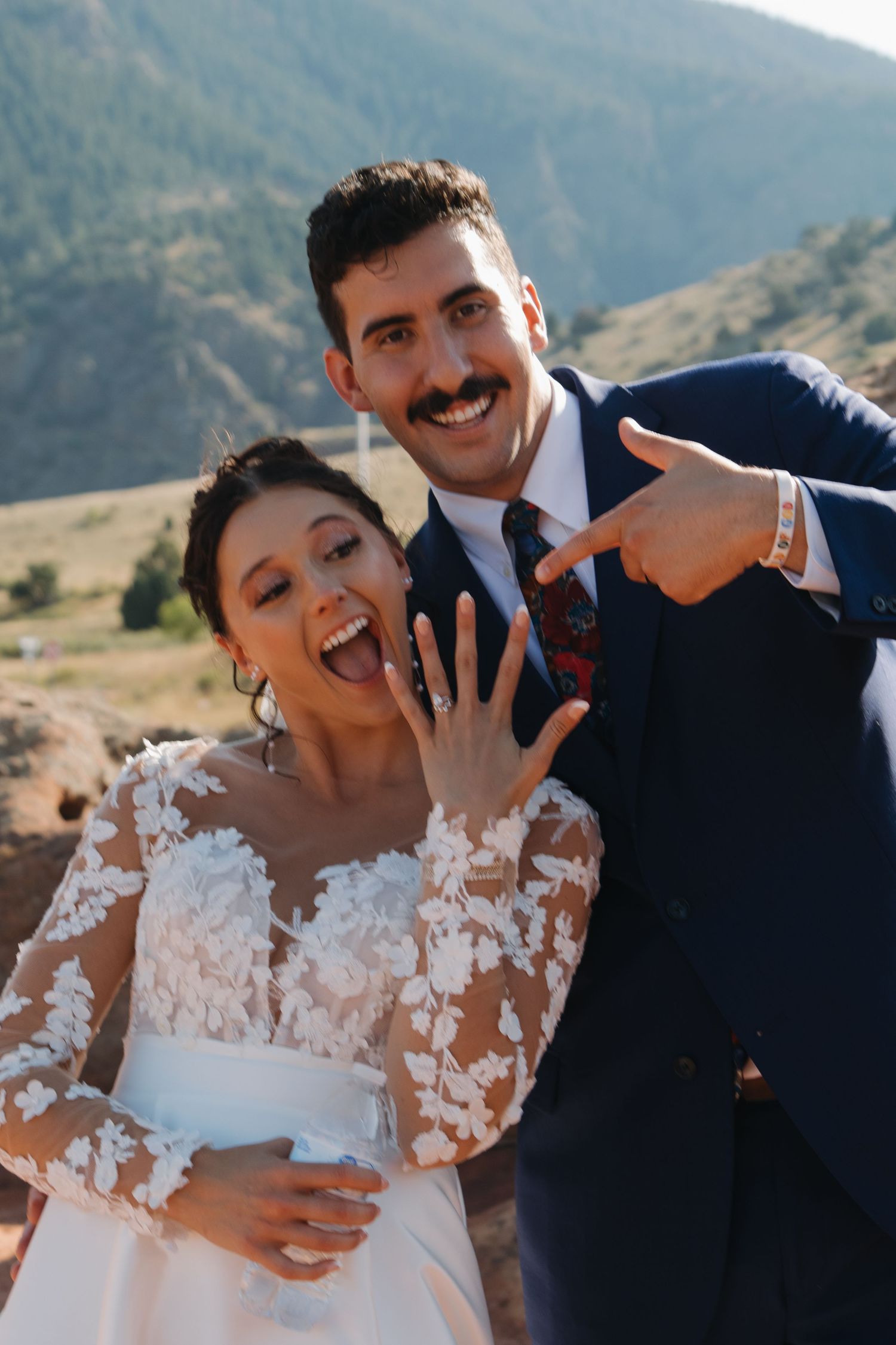 Newlyweds share joyful moment in front of mountain landscape wearing traditional wedding attire.