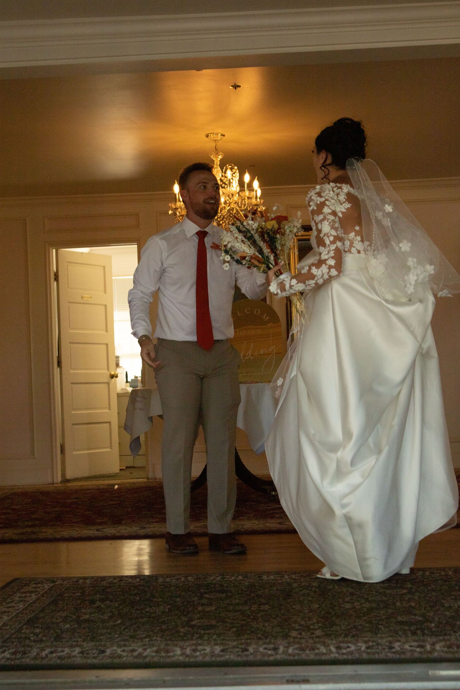 Wedding couple walking through elegant room with chandelier and carpeted floor.