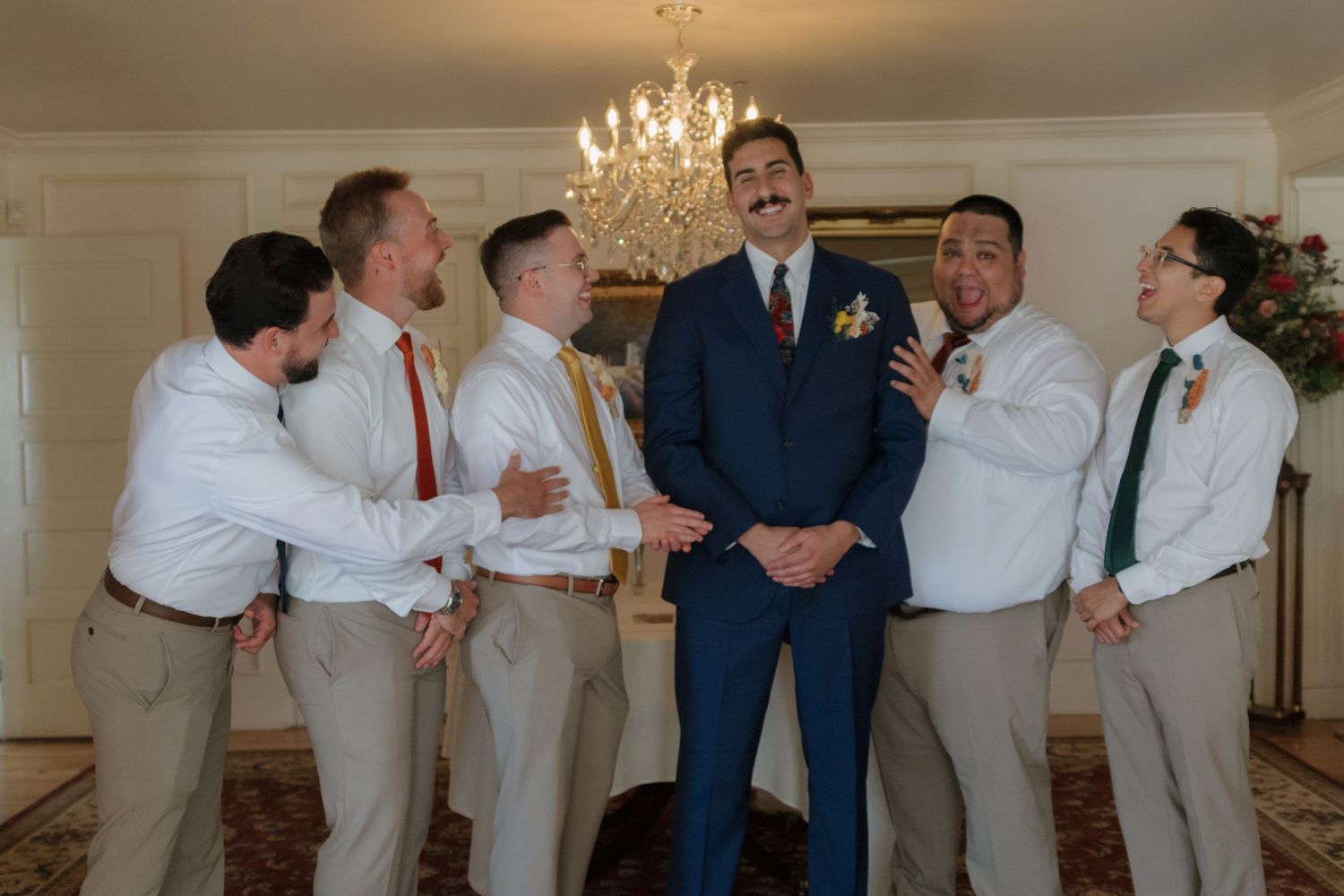 Groomsmen in blue suits and tan slacks posing together under a chandelier.