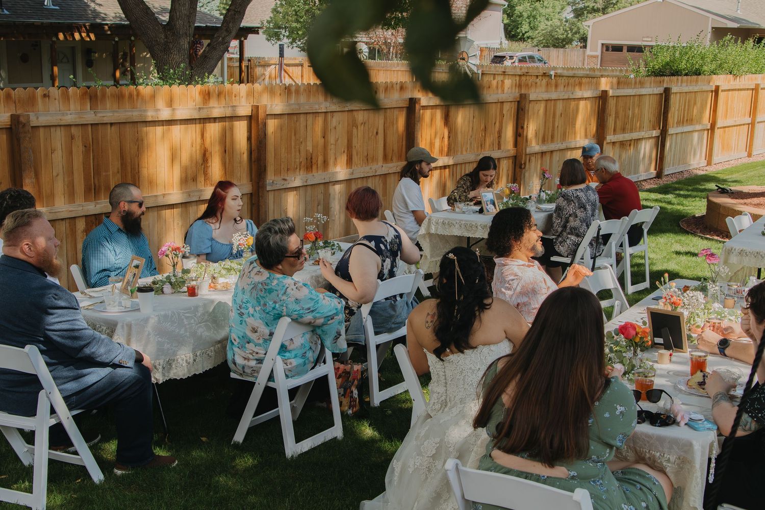 Outdoor wedding reception setup with white chairs and wooden fence in a backyard garden setting.