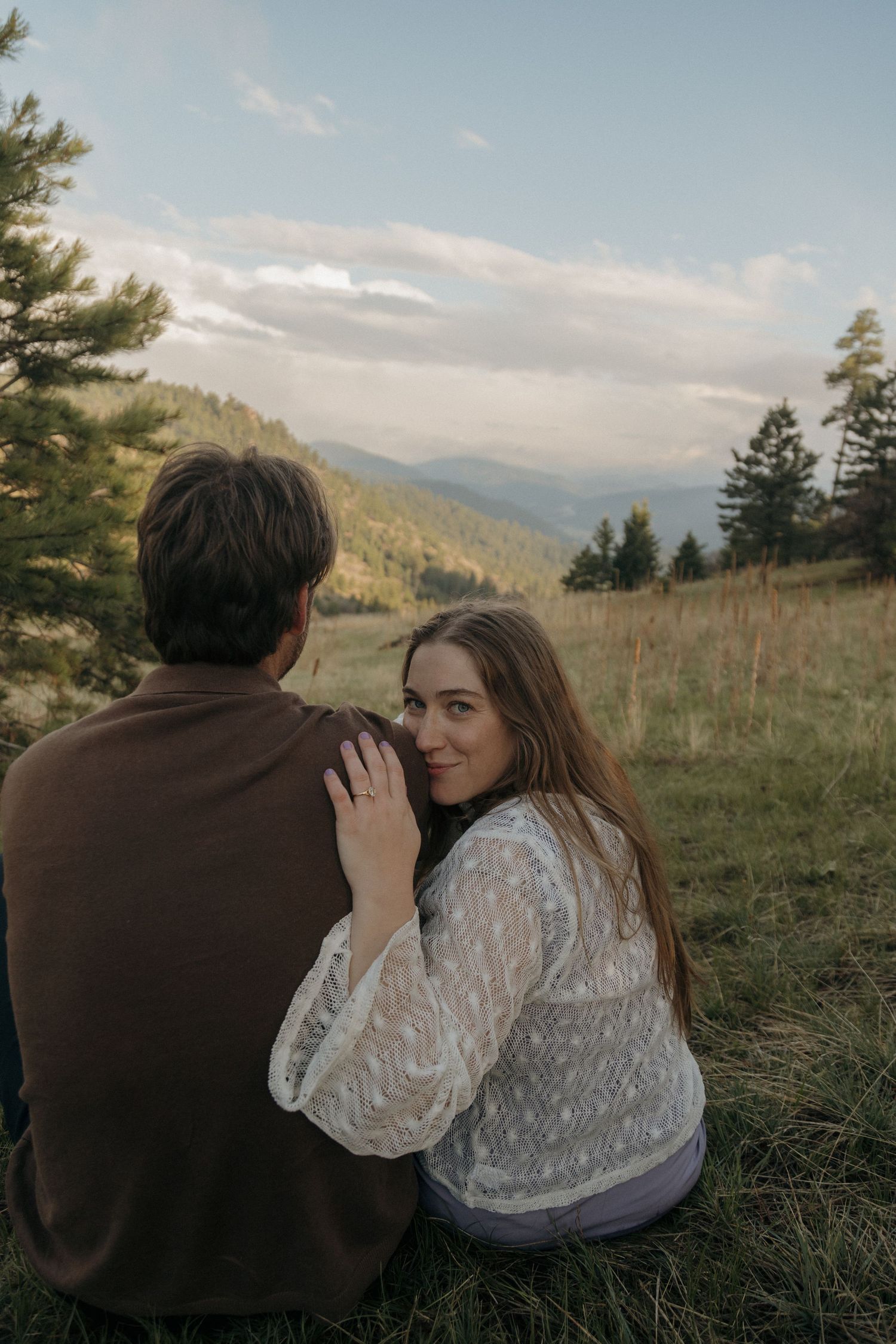 A couple sits on a grassy mountain meadow at sunset, woman in white lace blouse smiling over her shoulder.