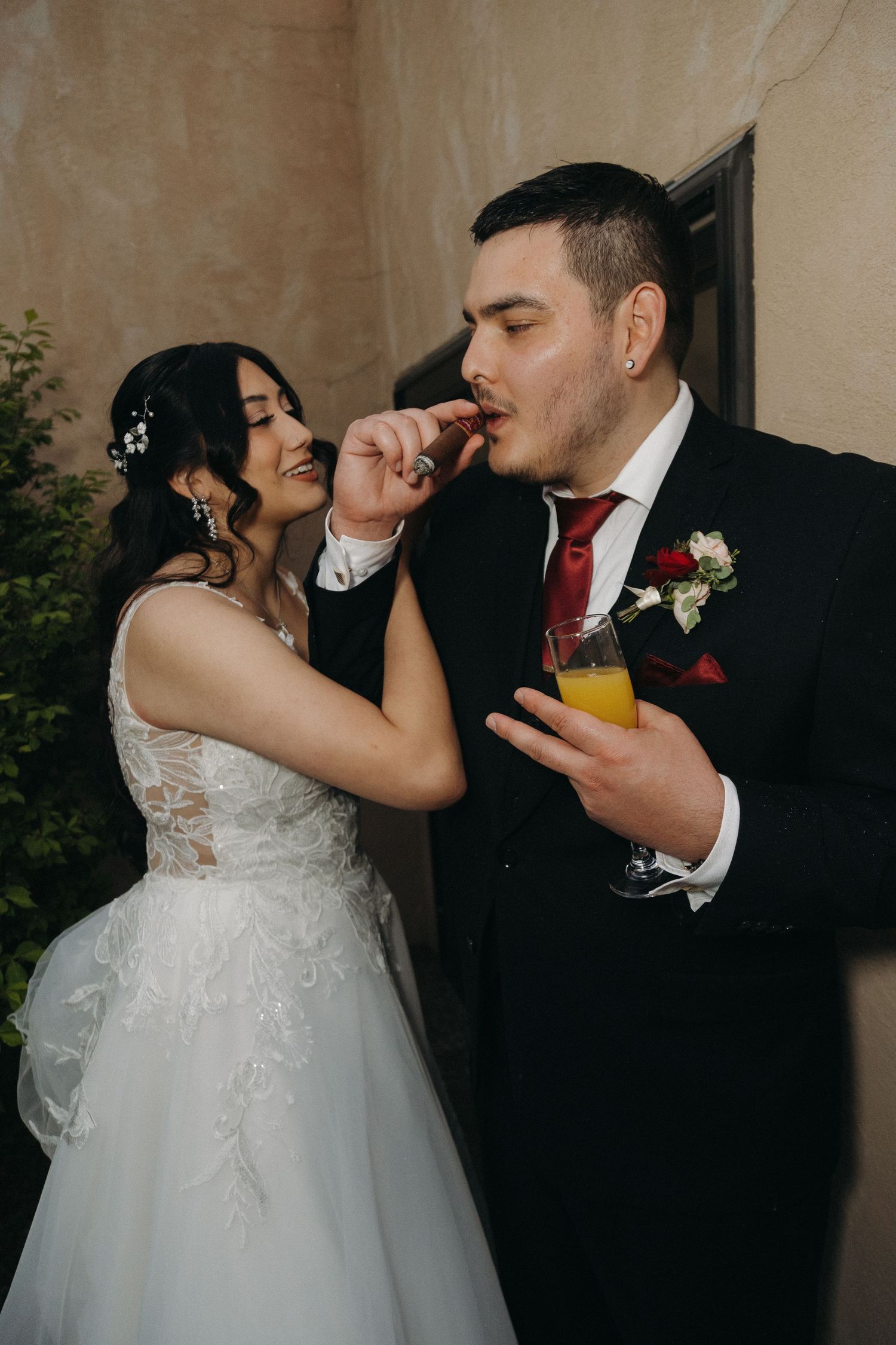 Bride feeding groom a cigar while he holds a glass of orange juice at their wedding reception.