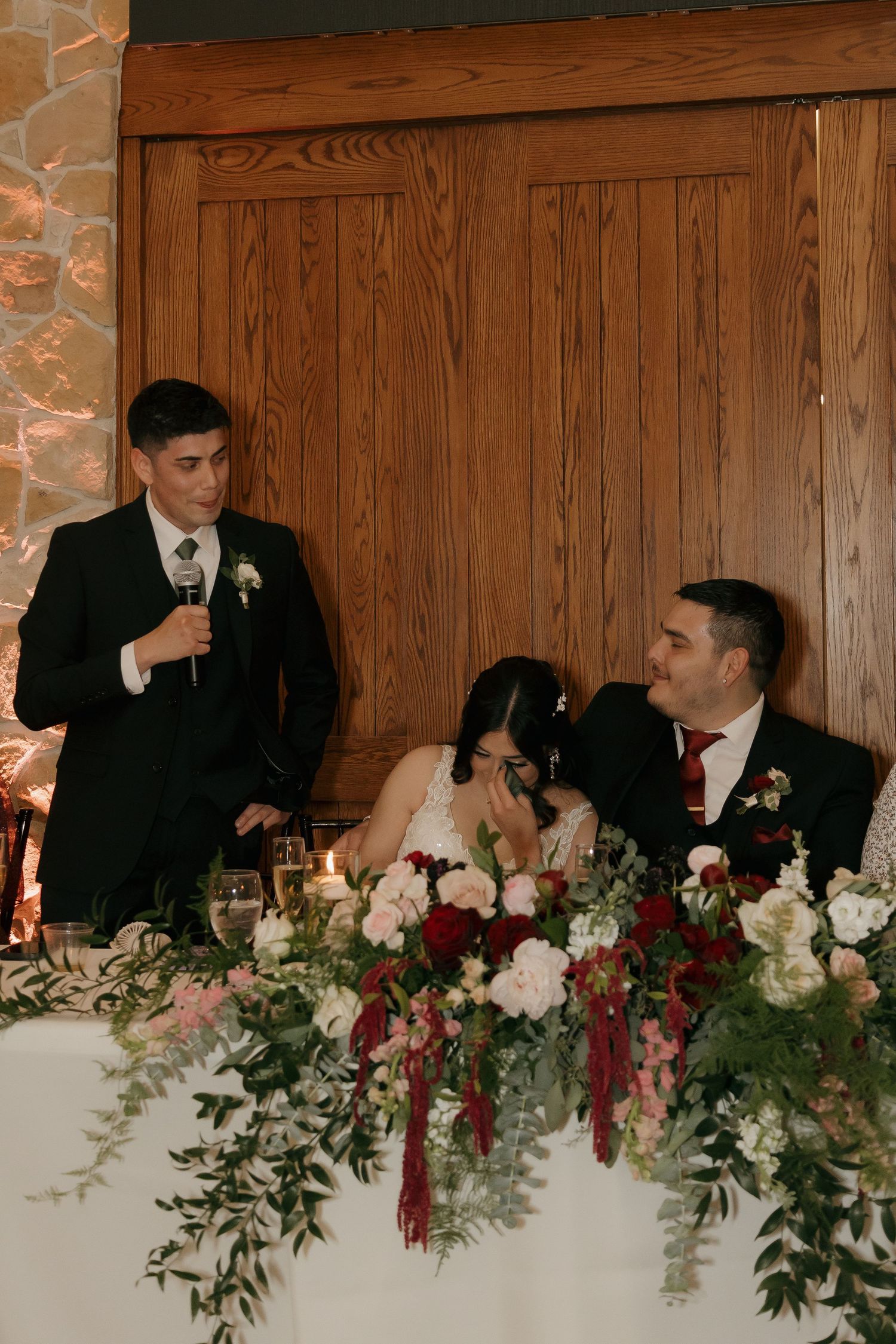 Best man gives a heartfelt speech at the wedding reception head table with floral arrangements.