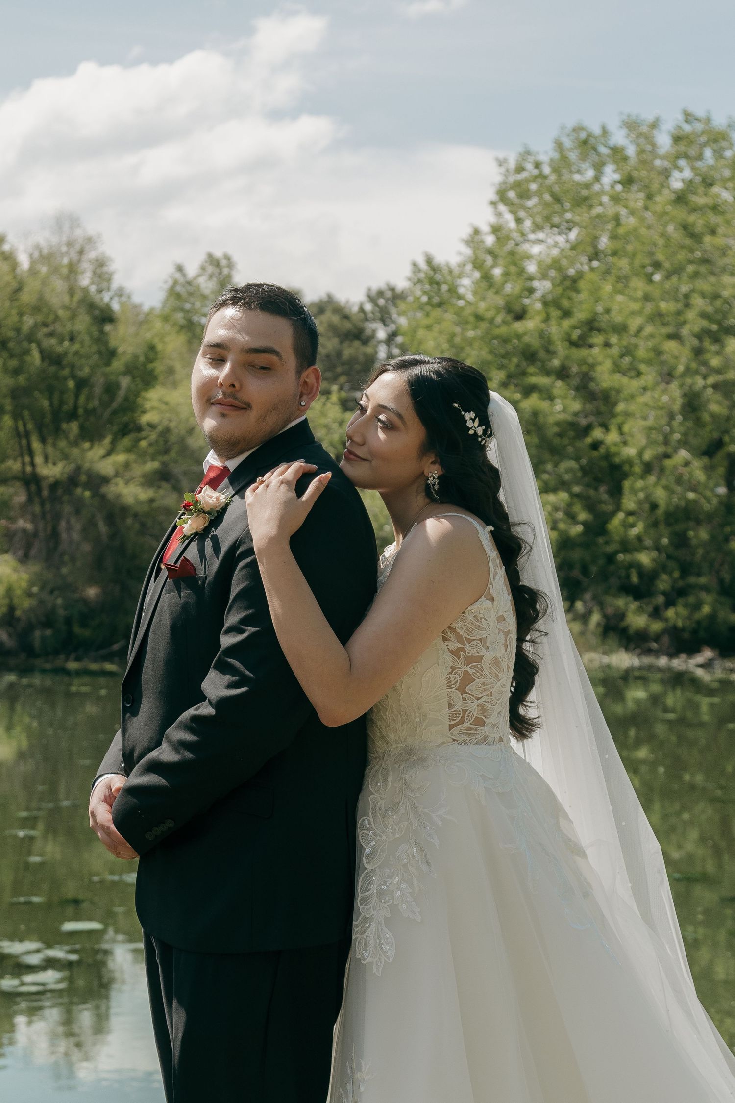 Bride in white lace gown holding groom's shoulder as they pose together by a serene lakeside backdrop.