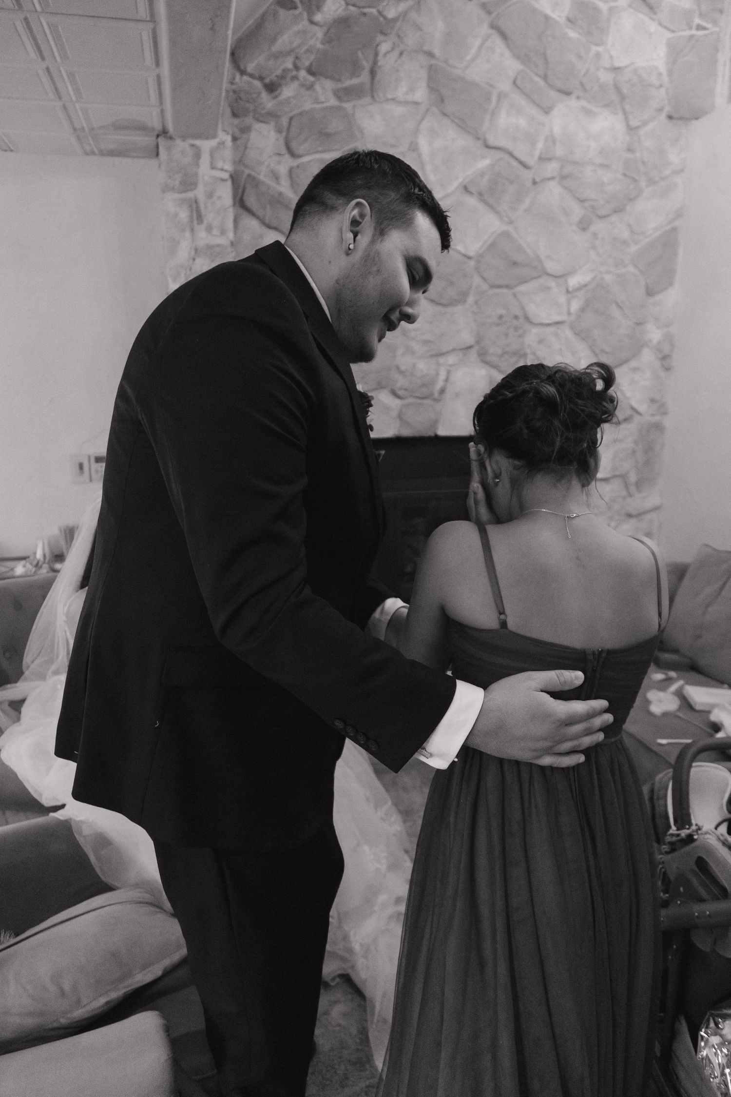 A groom in a black suit helps adjust a bridesmaid's green dress in a black and white photo.