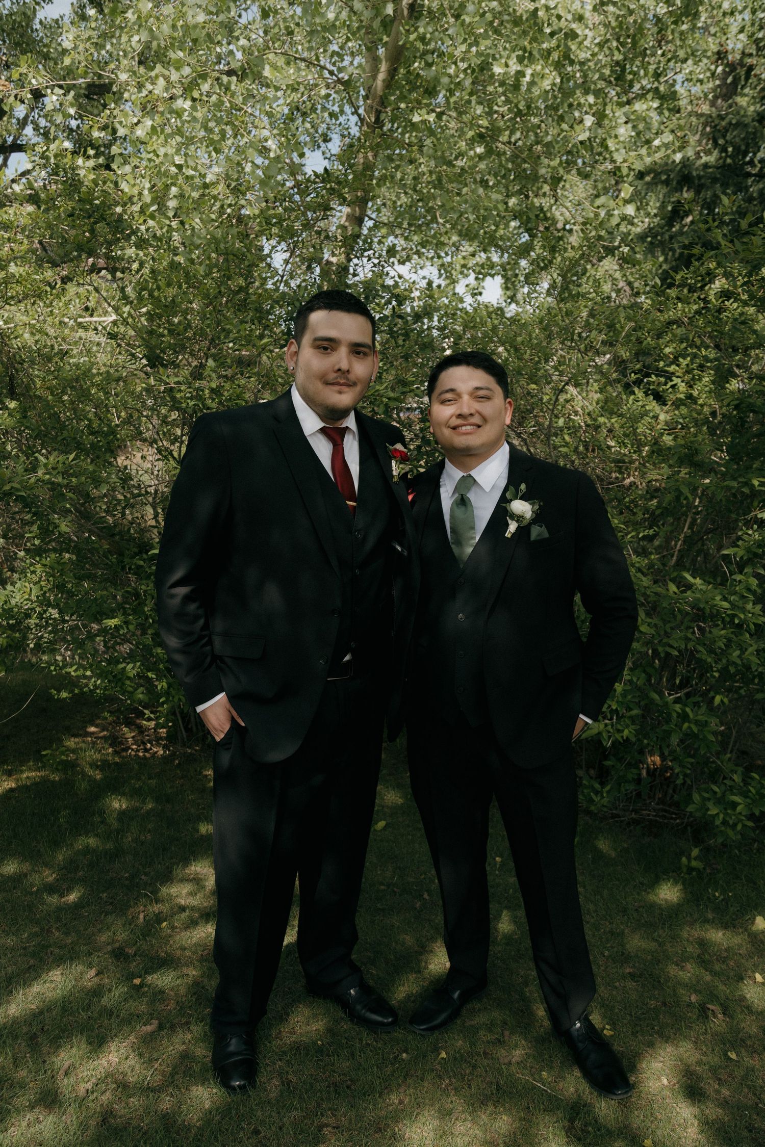 Two groomsmen in black suits with boutonnieres posing outdoors among green trees.