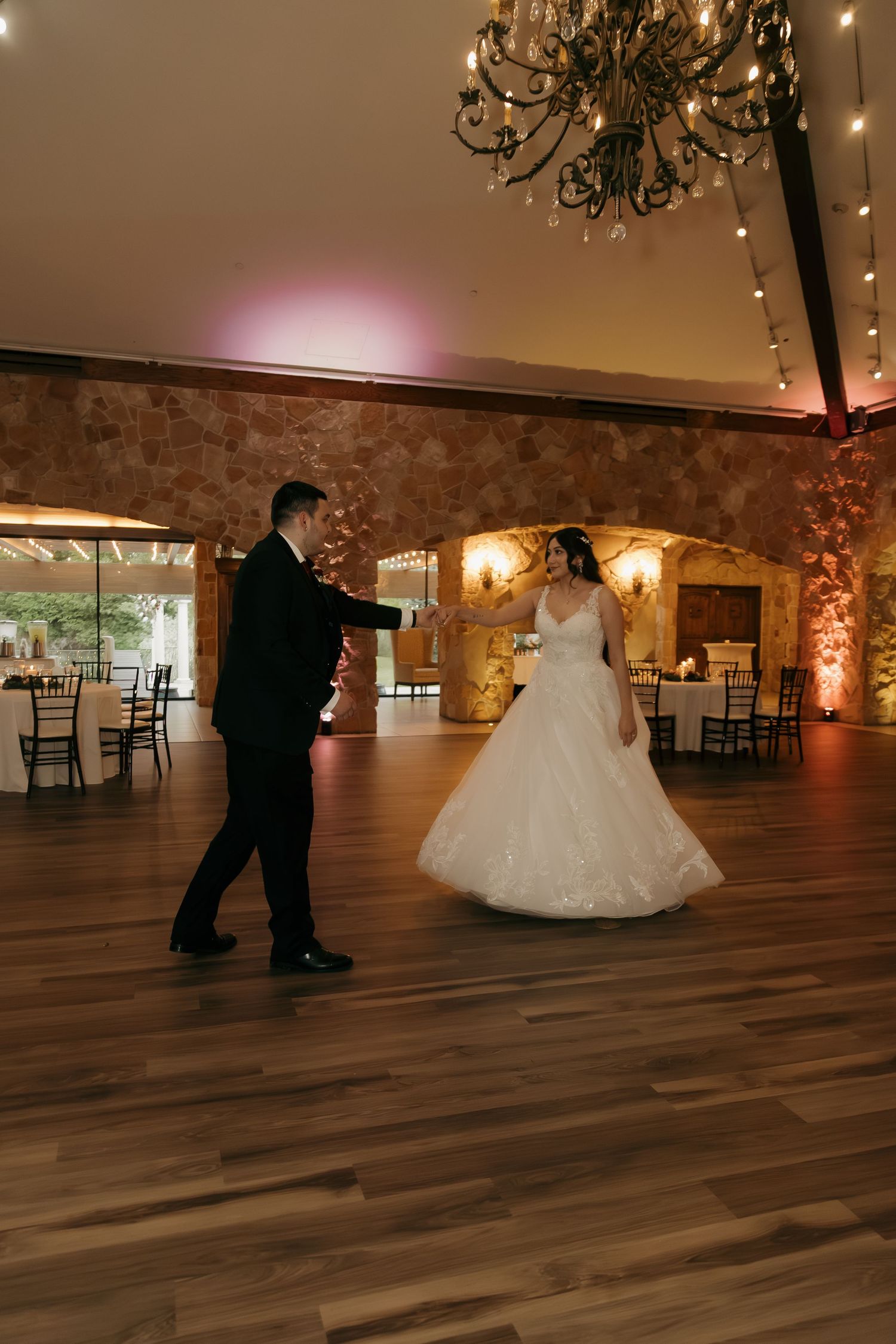 Bride and groom share romantic first dance under a chandelier in a warmly lit stone ballroom.