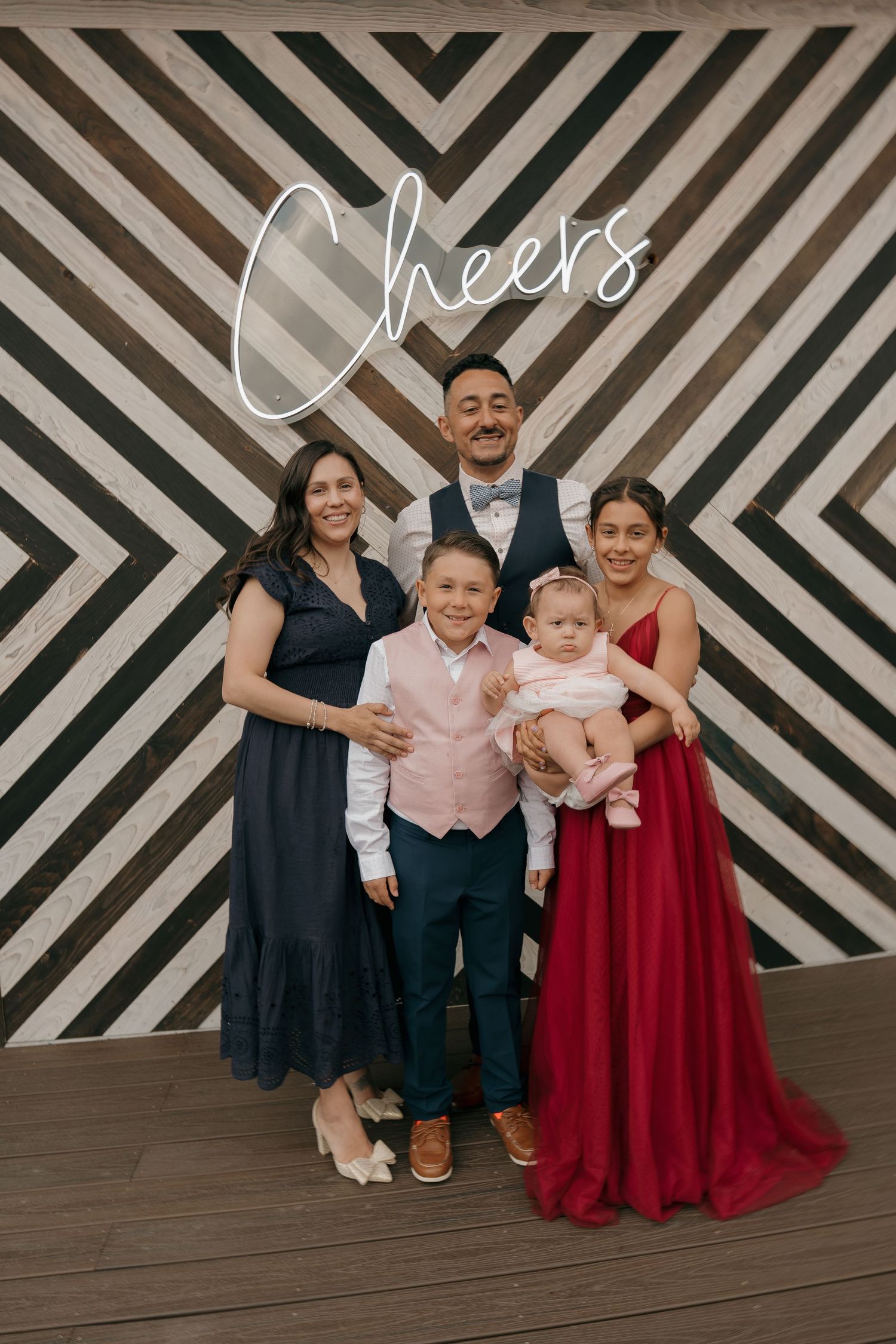 Family of five posing together at wedding reception in front of geometric chevron backdrop with Cheers sign.