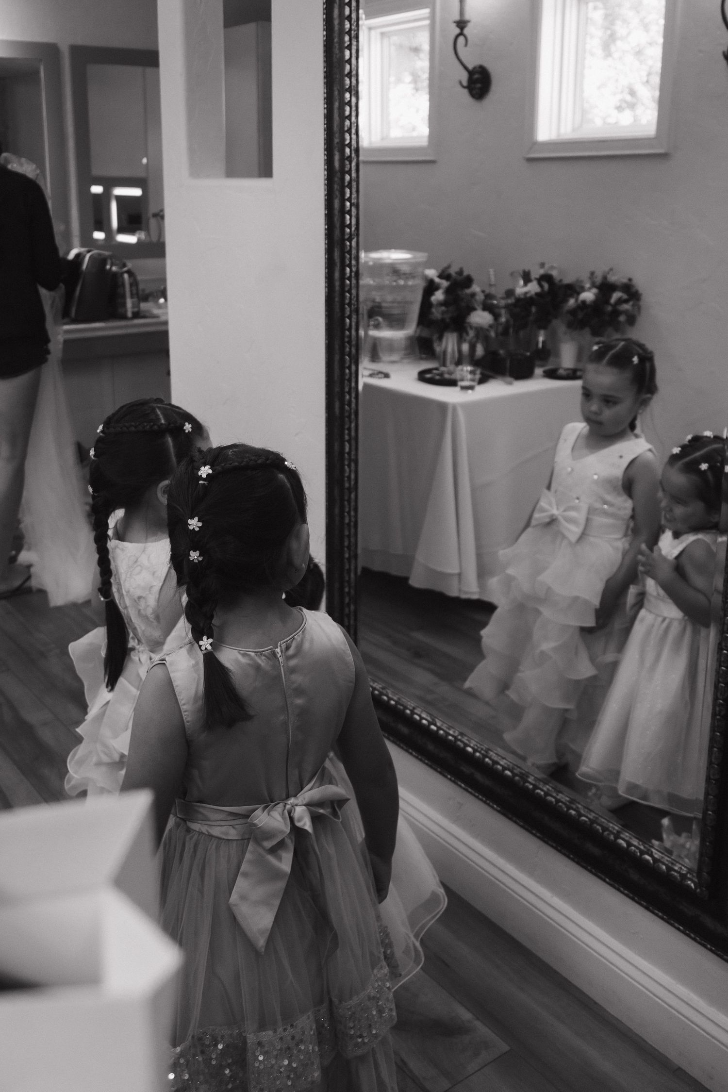 Black and white photo of a flower girl in a white dress looking at her reflection in a large mirror.