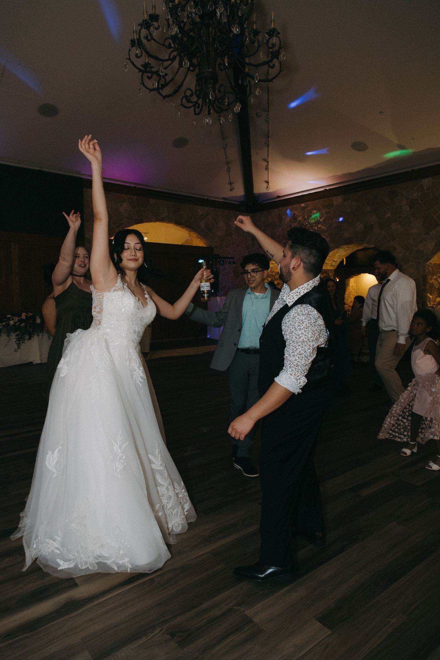 Bride in white lace gown dancing joyfully with groom on wedding reception dance floor surrounded by guests.