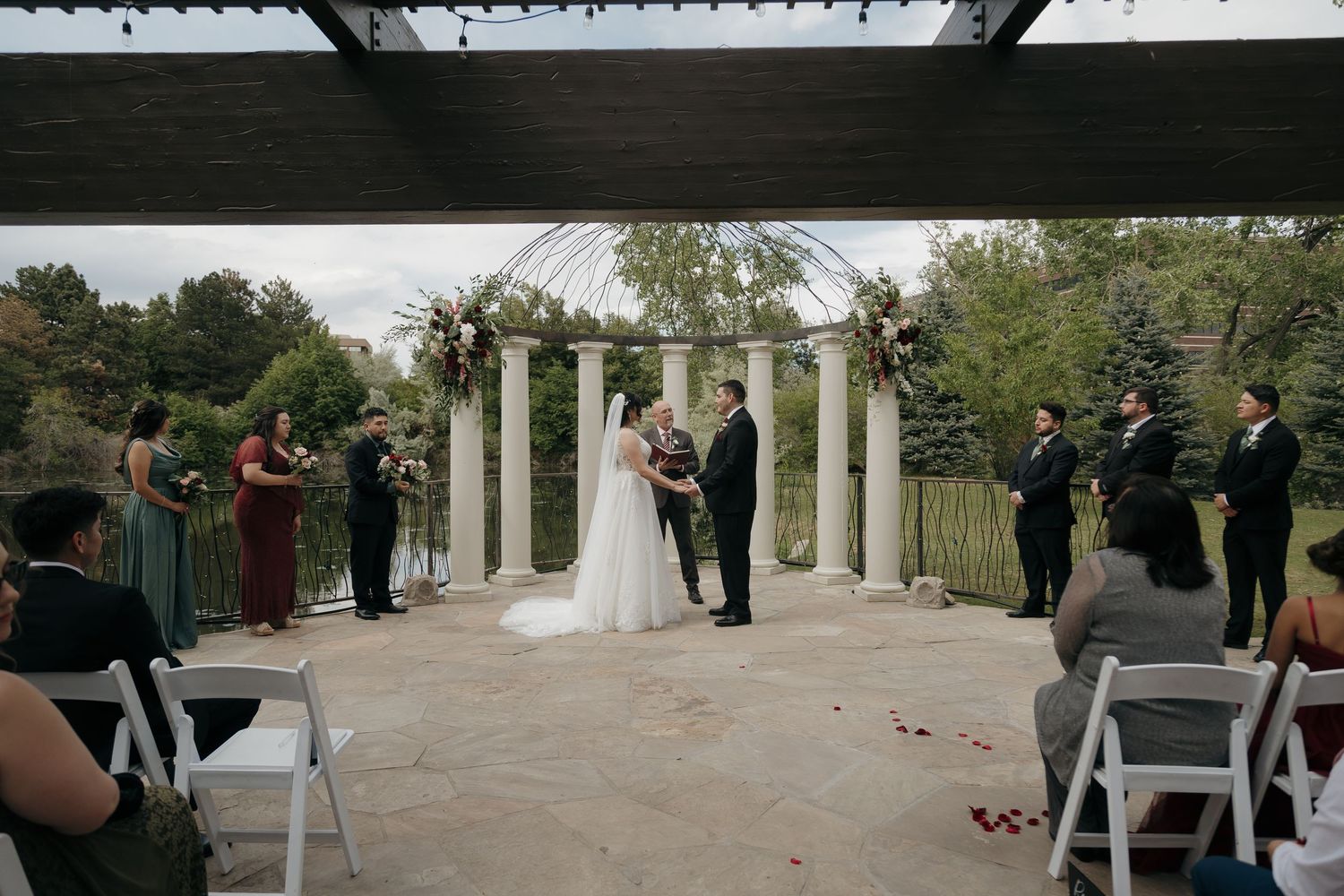 Bride and groom exchange vows at elegant outdoor wedding ceremony with white columns and floral arch.