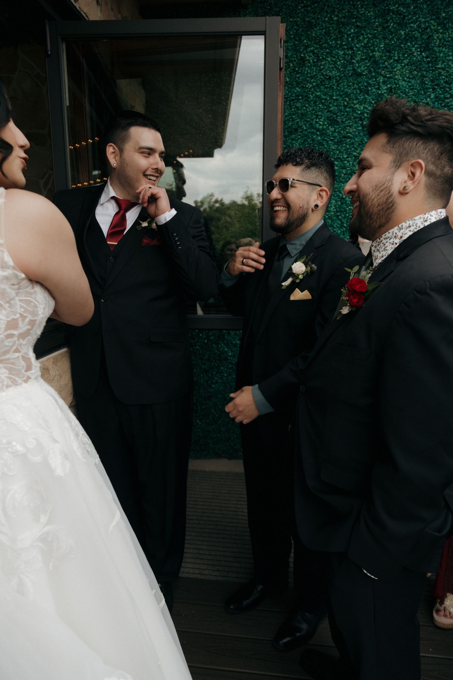 Groom and groomsmen in black suits laughing together in front of green hedge wall at wedding.