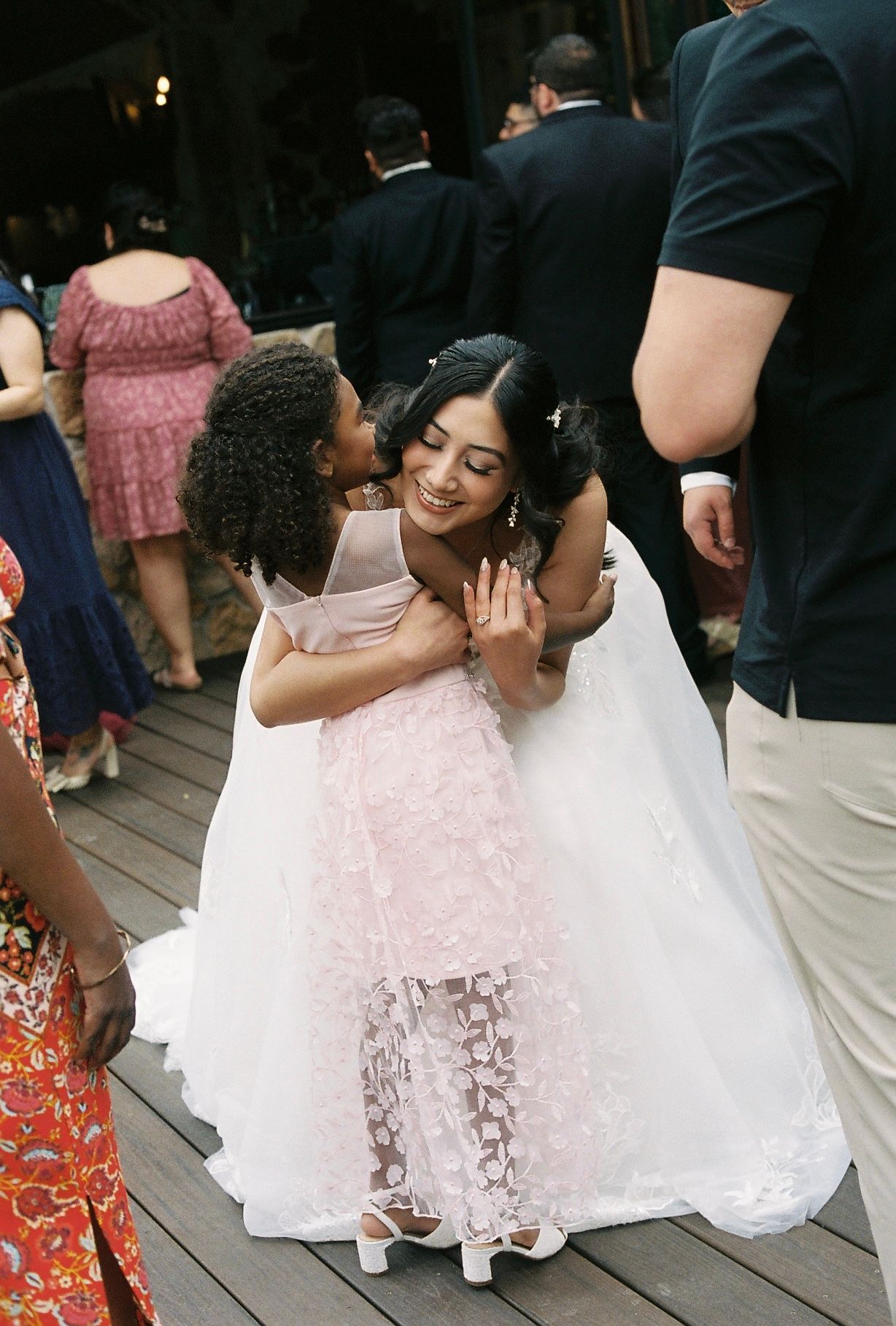 Bride in white wedding gown embraces young flower girl in pink dress on outdoor reception dance floor.