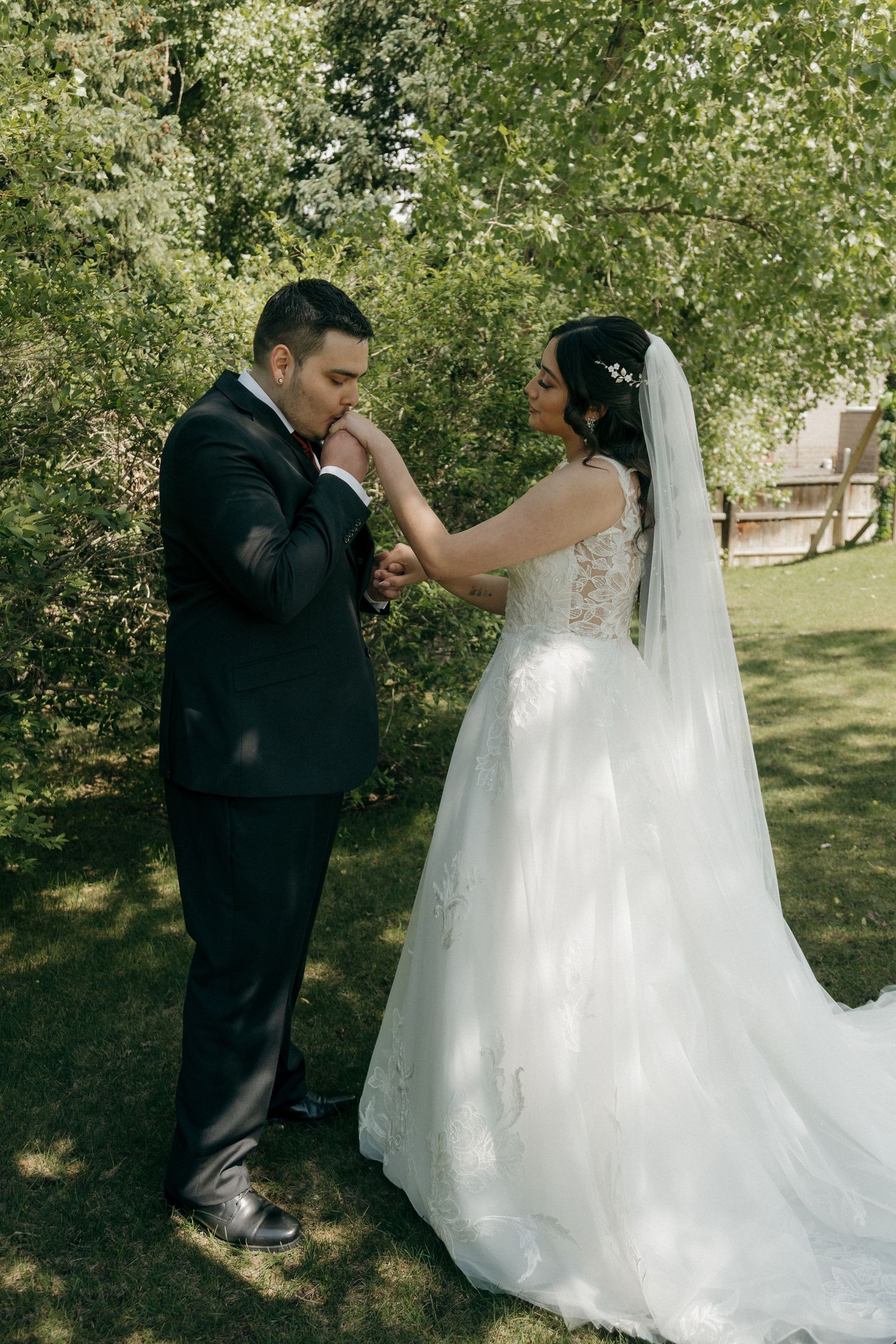 Groom gently kissing bride's hand during romantic outdoor first look moment surrounded by green trees.