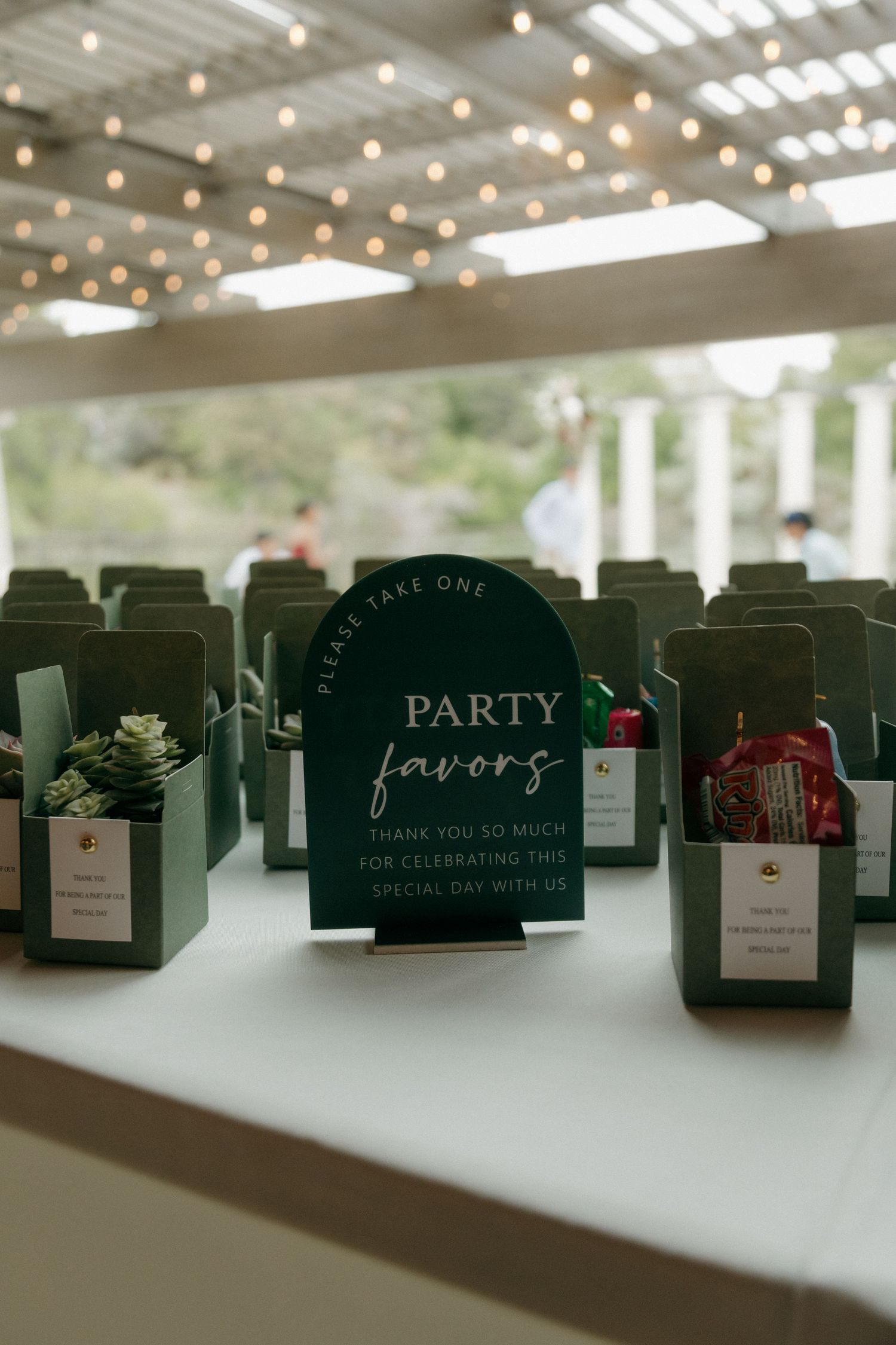Wedding party favors displayed in green boxes with a sign on a white table under string lights.