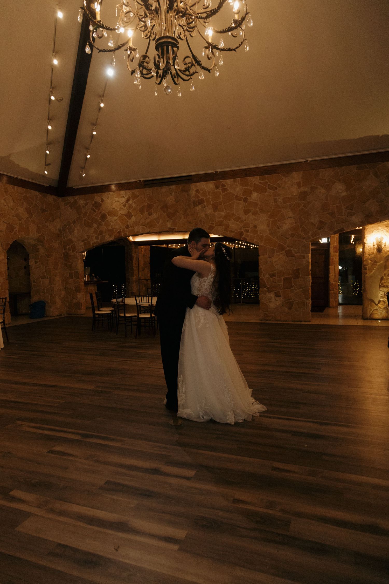 Bride and groom share an intimate first dance under a chandelier in a romantic stone-walled wedding venue.