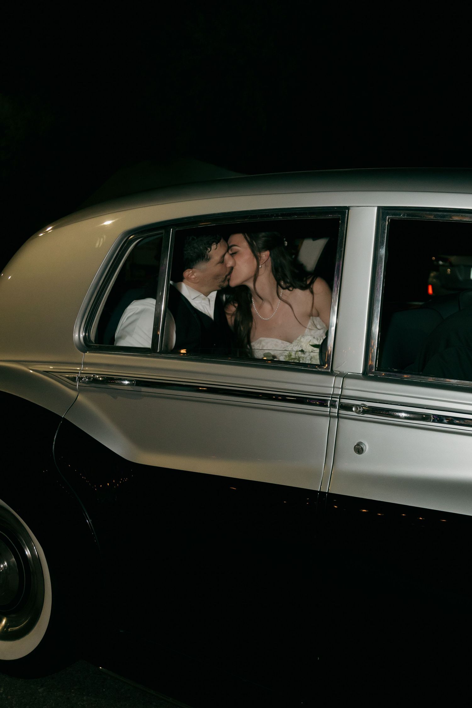 Newlywed couple sharing romantic kiss inside vintage classic car after wedding reception.